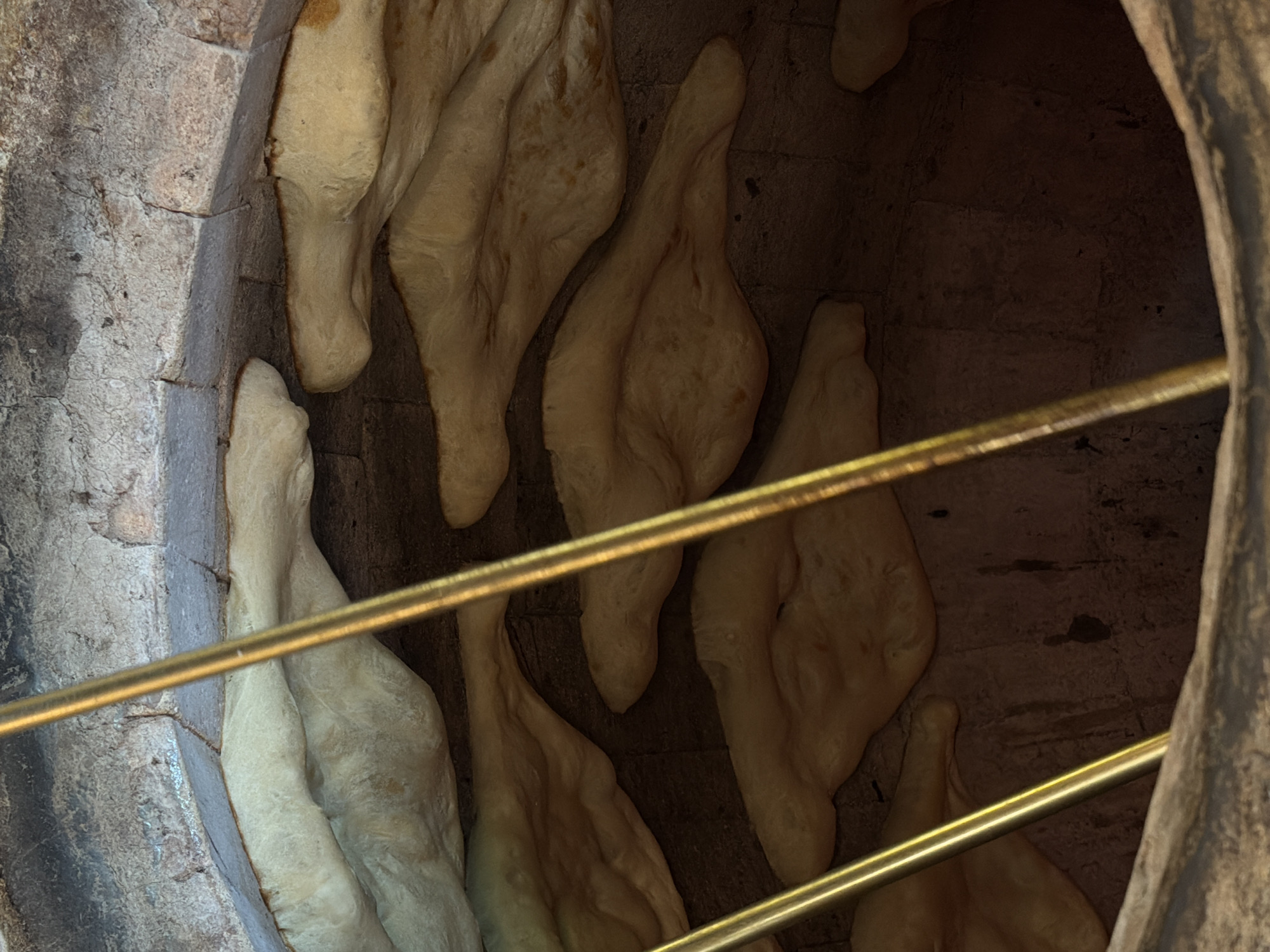 The image captures the interior of a traditional oven, likely a *tonir*, used for baking bread, located in Tsovagyugh, Armenia. Several pieces of light-colored dough, appearing to be in various stages of baking, are adhered directly to the rough, dark, brick or stone walls of the cylindrical oven. The dough pieces are irregularly shaped, long, and somewhat flattened, with some areas showing a slightly browner, cooked texture.
Two shiny, golden metal bars extend horizontally across the frame, positioned in the foreground, partially obscuring the view into the oven. These bars might serve as a protective barrier or a structural element for the baking process. The interior of the oven is dimly lit, emphasizing the rustic textures of the stone walls and the dough. No people are visible, and the scene focuses on the traditional method of baking bread. There is no visible text in the image.