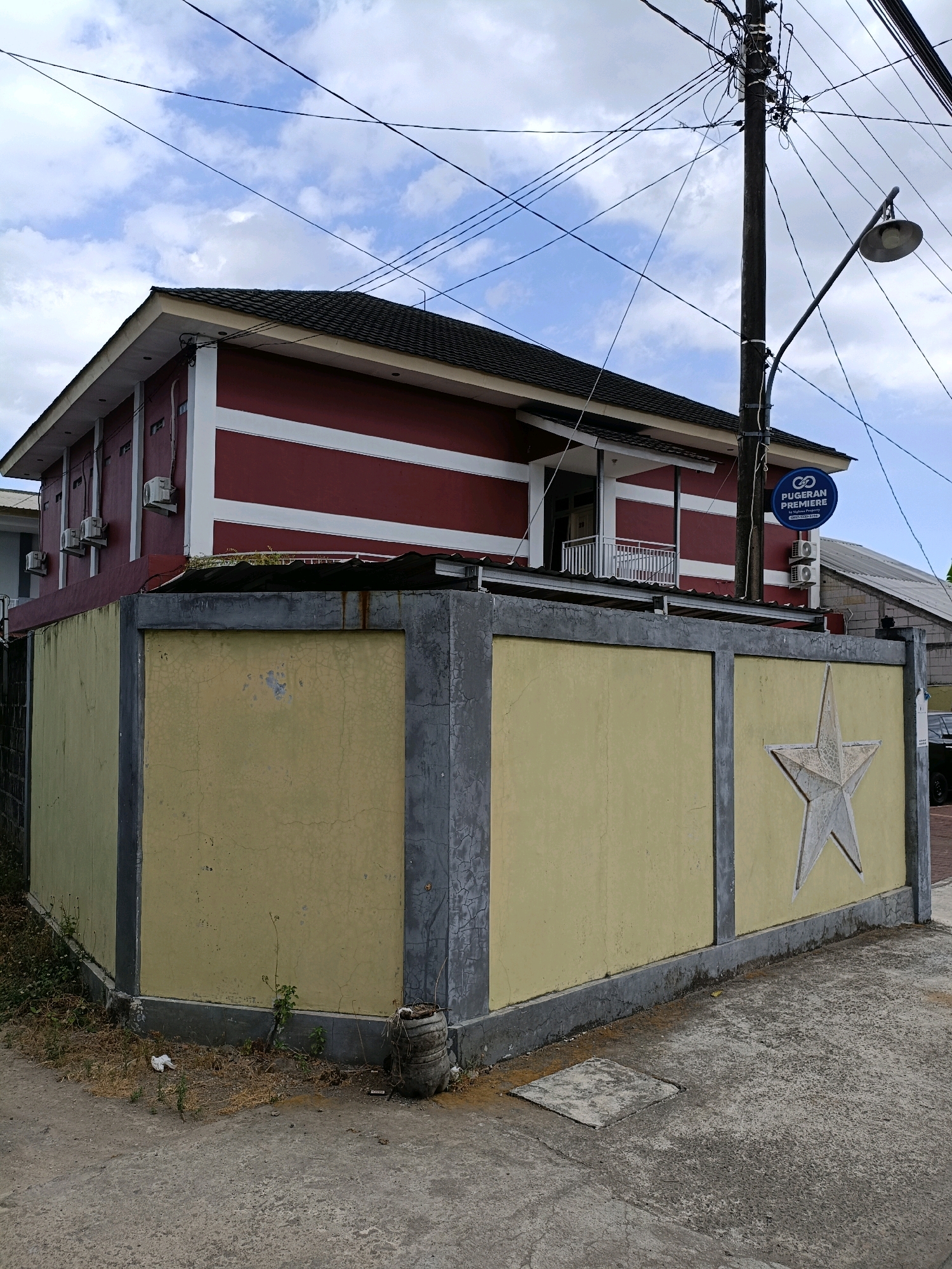The image shows a building with a red and white striped facade, located in Maguwoharjo, Indonesia. The building has multiple air conditioning units visible on its exterior. In front of the building is a low, yellow wall with a large, gray, raised star emblem on its surface. The wall is bordered by concrete pillars. A lamppost with a visible sign that reads "PUGERAN PREMIERE" is positioned near the wall. The sky is partially cloudy. The ground in front of the wall is paved with concrete, with some dry vegetation and debris. The scene appears to be a street-level view in a somewhat commercial or residential area.