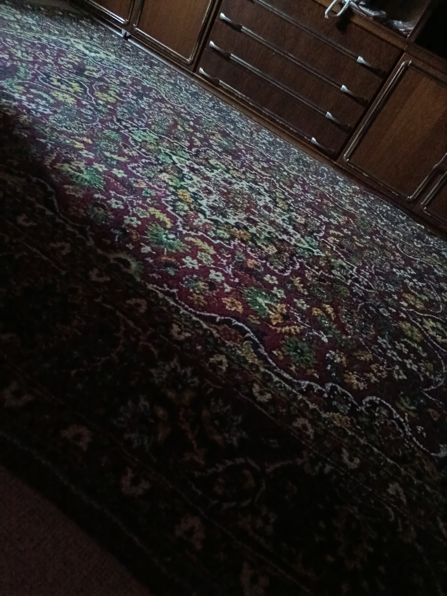 The image shows a close-up, angled view of a richly patterned carpet. The carpet has a deep red base with intricate floral and paisley motifs in colors including yellow, green, and white. The lighting is dim, casting some areas into deep shadow, particularly towards the bottom of the frame, making the details of the carpet less discernible in those sections. In the upper right portion of the image, a dark wooden dresser with multiple drawers is visible. The dresser has metallic drawer pulls and appears to be a piece of furniture within a room. The overall impression is one of a domestic interior. There is no visible indication of the city of Ekibastuz, Kazakhstan, nor any people or actions taking place.