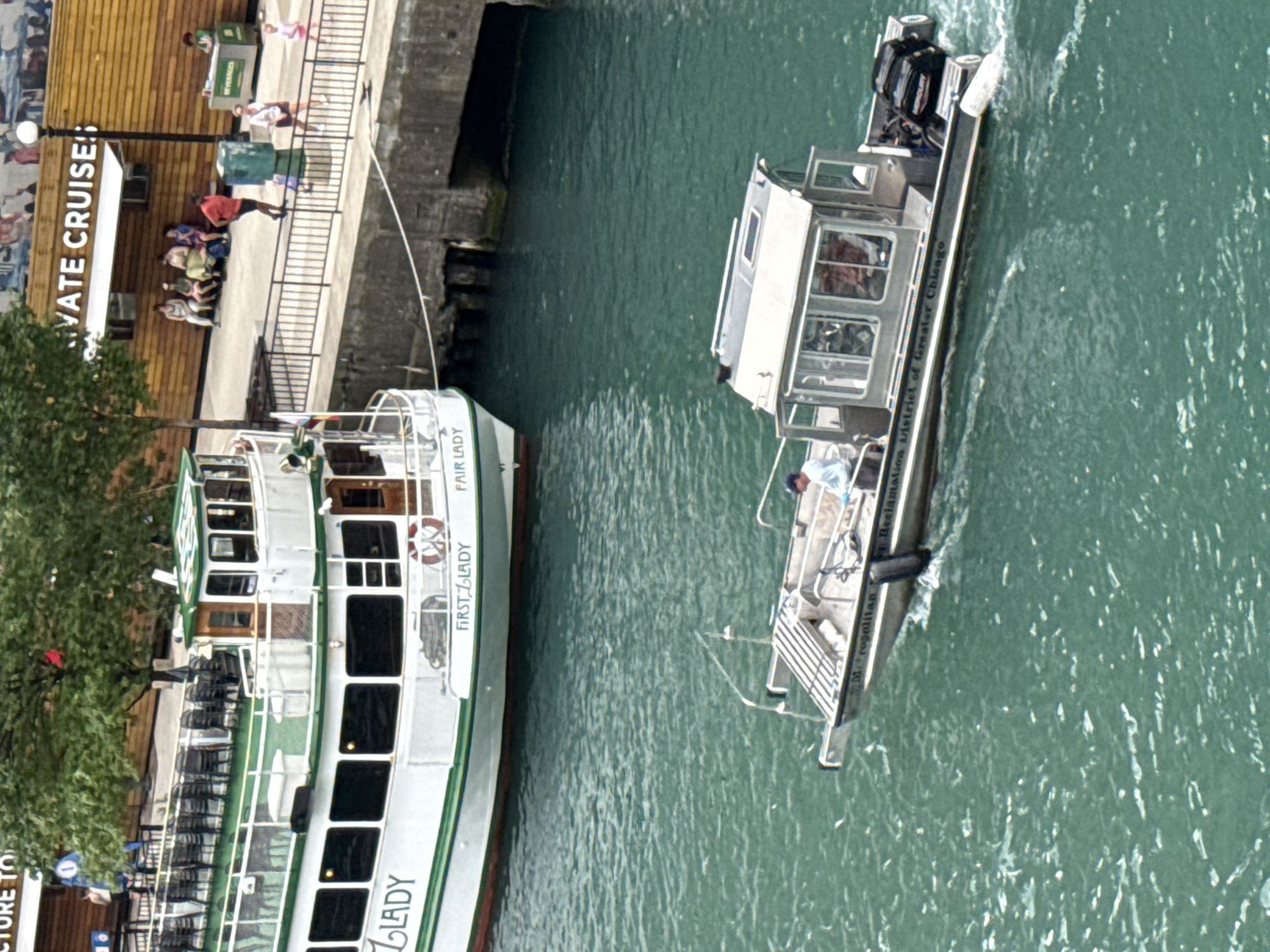 This is an aerial medium shot of a boat in the Chicago River, Chicago, Illinois, United States of America. On the left, a large white passenger boat with green trim, named "LADY", is docked. The passenger boat has several levels and many windows. The name "FIRST LADY" and "FAIR LADY" are visible on its side. On the right, a smaller, gray boat with a cabin is moving in the river. The boat has "The Metropolitan District of Greater Chicago" written on its side. A person in a light-colored shirt and dark cap is at the helm of the smaller boat. In the background, a wooden building with the words "IVATE CRUISES" is visible. People are walking on the pier next to the boats. The water is a turquoise color.