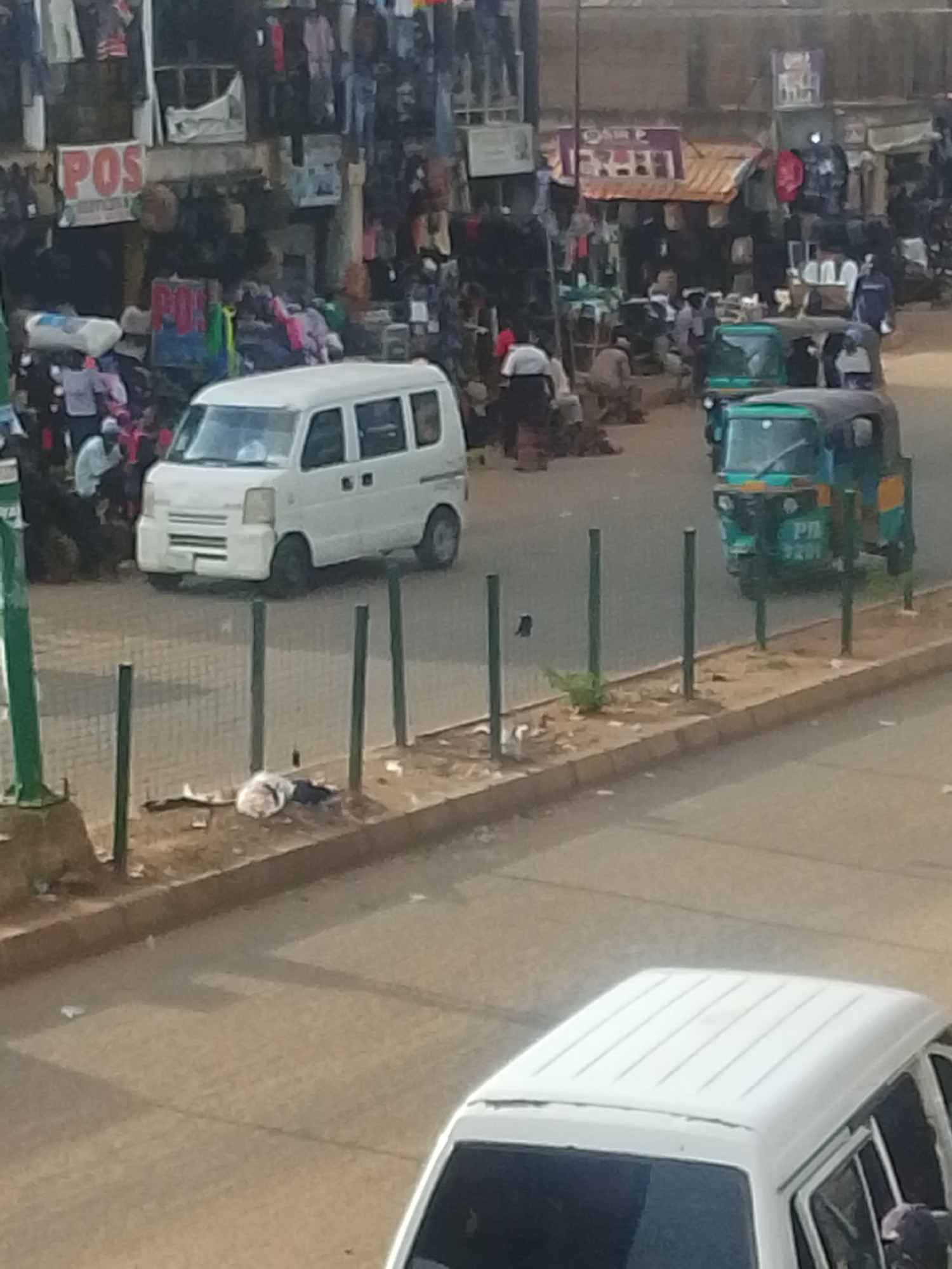 The image depicts a street scene in Bukuru, Nigeria, characterized by a bustling marketplace environment. The foreground shows a street with a median barrier of green posts. A white car is partially visible in the bottom of the frame. Adjacent to the road is what appears to be litter, including possibly discarded clothing. In the middle ground, a white minivan is parked near the edge of the road, and two "keke napeps", which are three-wheeled vehicles. In the background, market activity is apparent with several figures interacting amidst stalls displaying clothing and other goods. Signage reading "POS" is seen. People are moving and engaging in buying/selling transactions. The area is congested with vehicles and pedestrians.