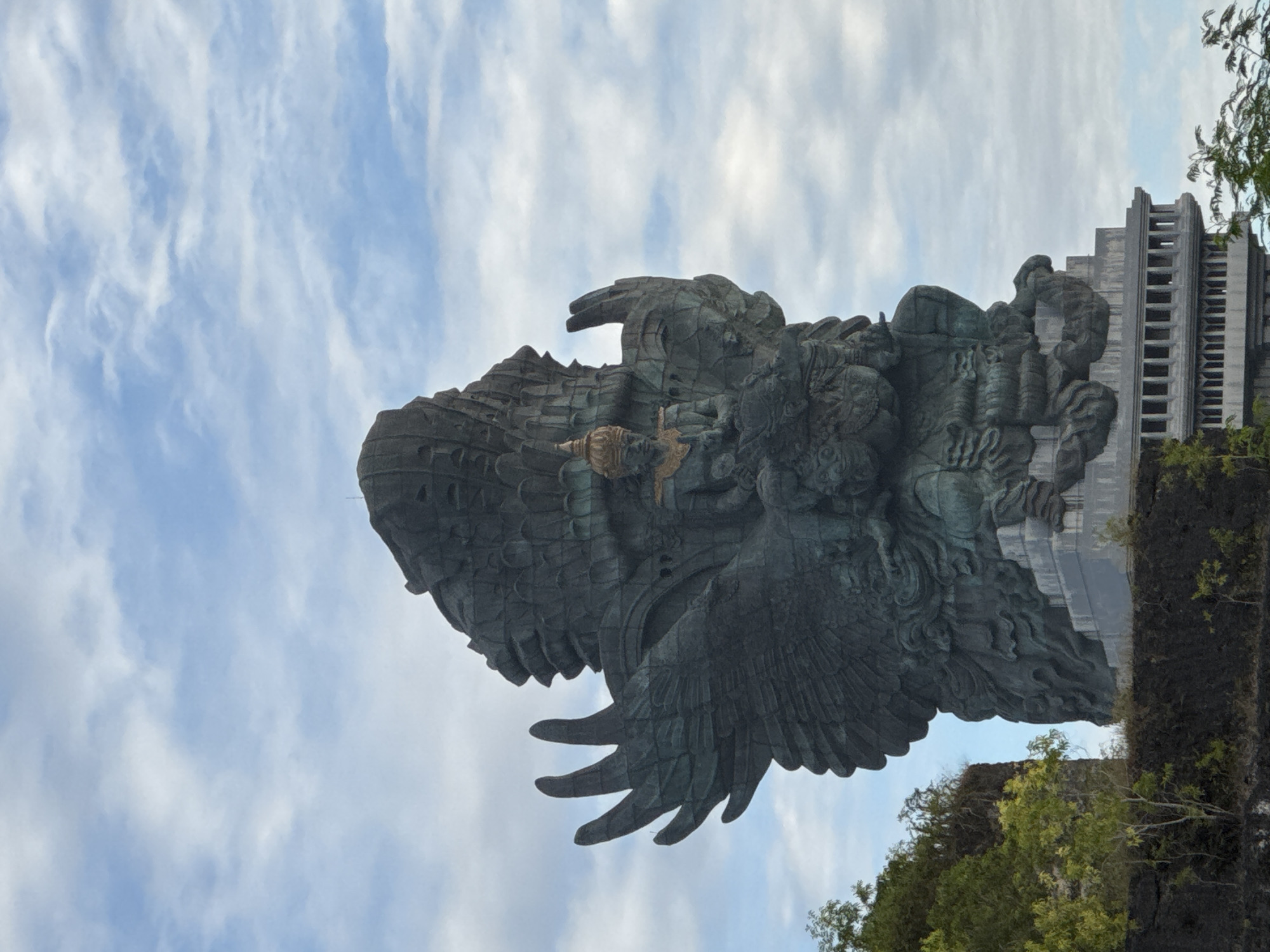 The image shows the Garuda Wisnu Kencana statue located in Kuta Selatan, Indonesia. The monument features a large, copper and brass sculpture of the Hindu god Vishnu riding on the mythical bird Garuda. The statue dominates the scene and is positioned against a backdrop of a cloudy sky. The base of the statue includes a building with multiple floors. The lower part of the scene is covered with greenery.