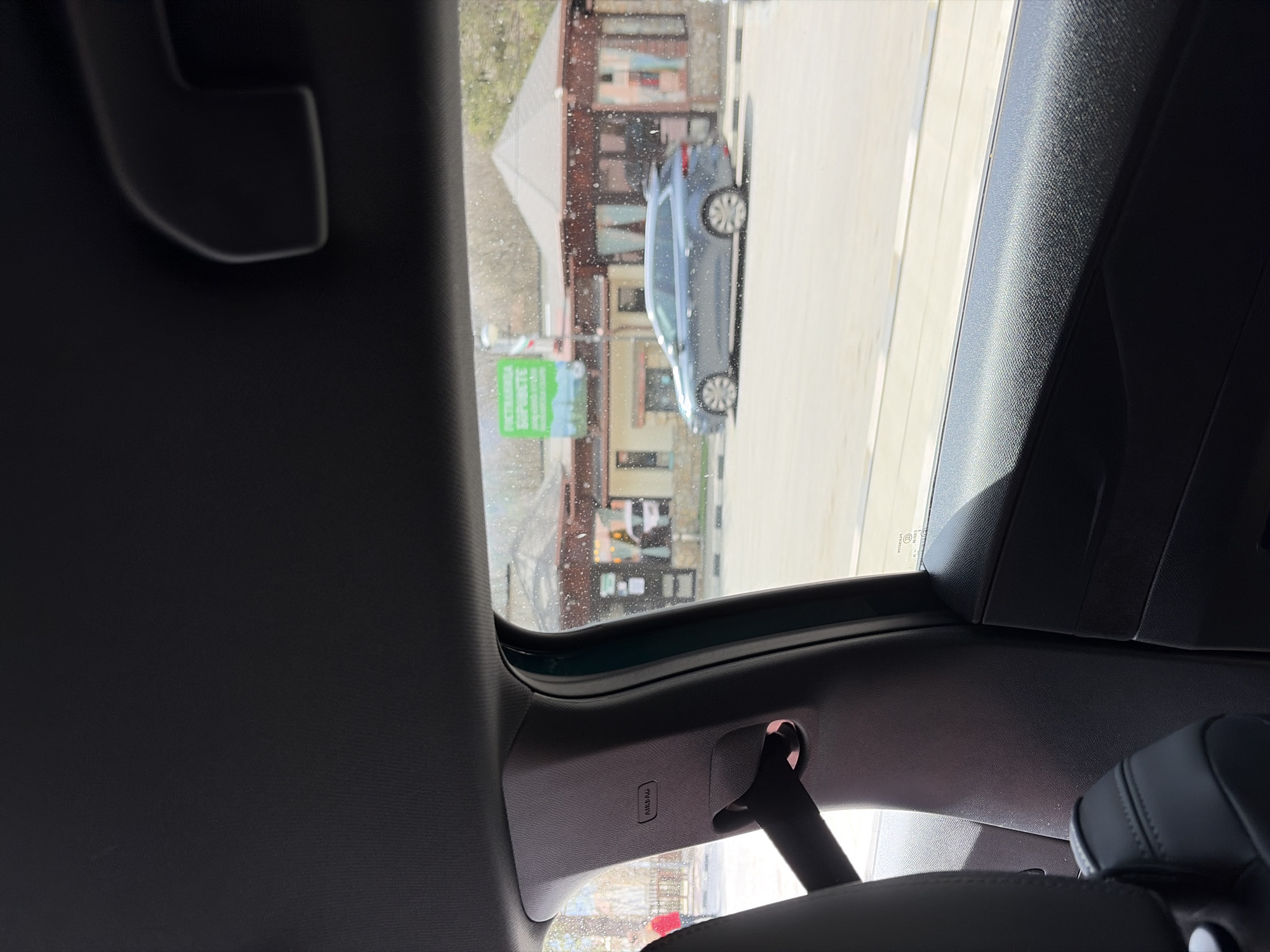 The image depicts an interior view from a car in Gintsi, Bulgaria. The car's sunroof is open, revealing a street scene. A parked light blue car is visible outside. The street has buildings on the side. The car's interior includes the headrest of a seat, a seatbelt attachment, and the dark-colored roof and panels of the car. The environment is typical of a small Bulgarian town. There are no people visible.