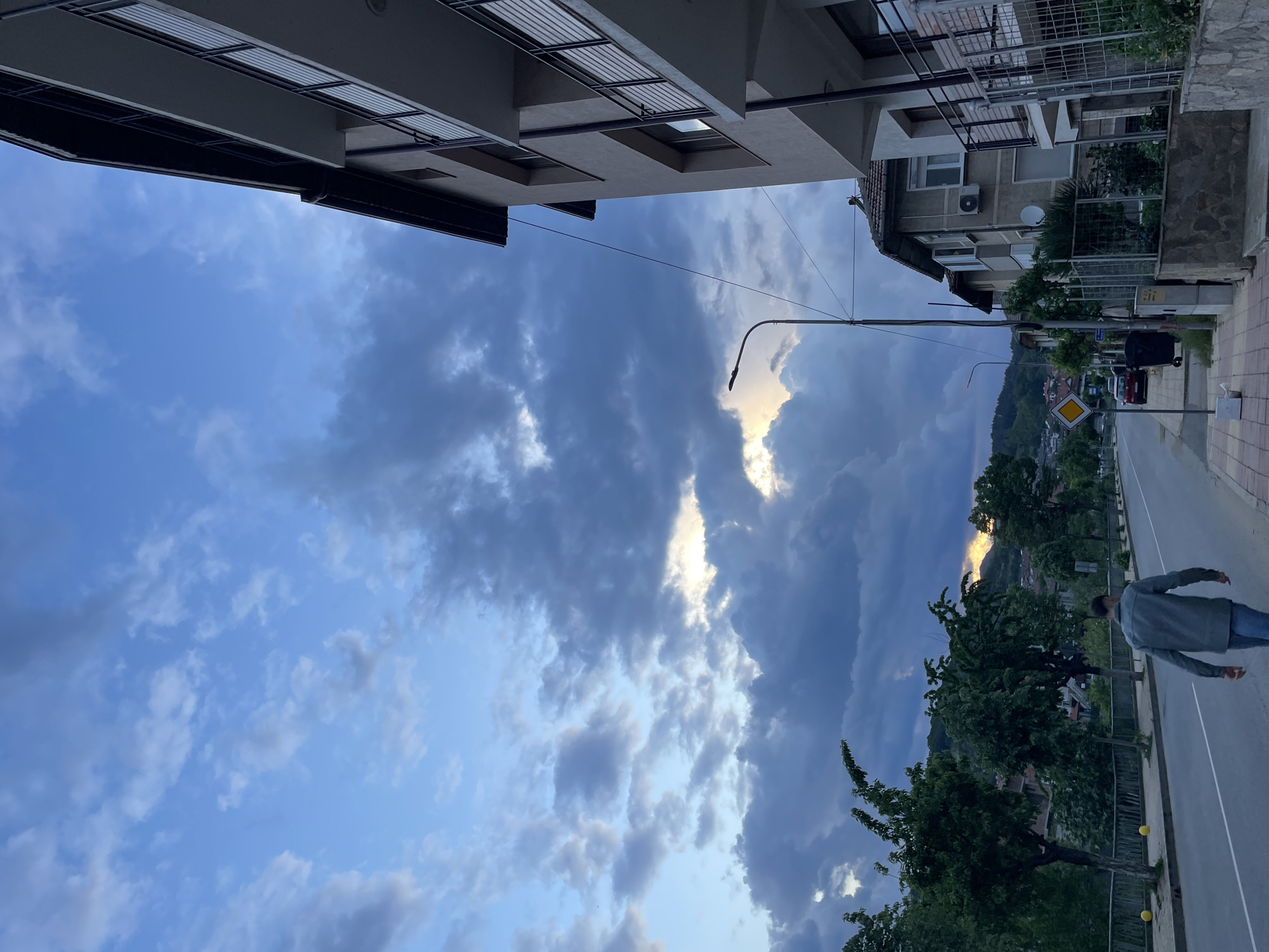 The image shows a street scene in Zlatograd, Bulgaria, taken during the evening. The sky is partially cloudy with light shining through. Buildings frame the view from above. A street with a sidewalk runs alongside. A person dressed in a gray jacket and blue jeans walks on the road. Trees and greenery are visible on the sides of the street. A street light and a road sign are also present. In the distance, mountains can be seen.
