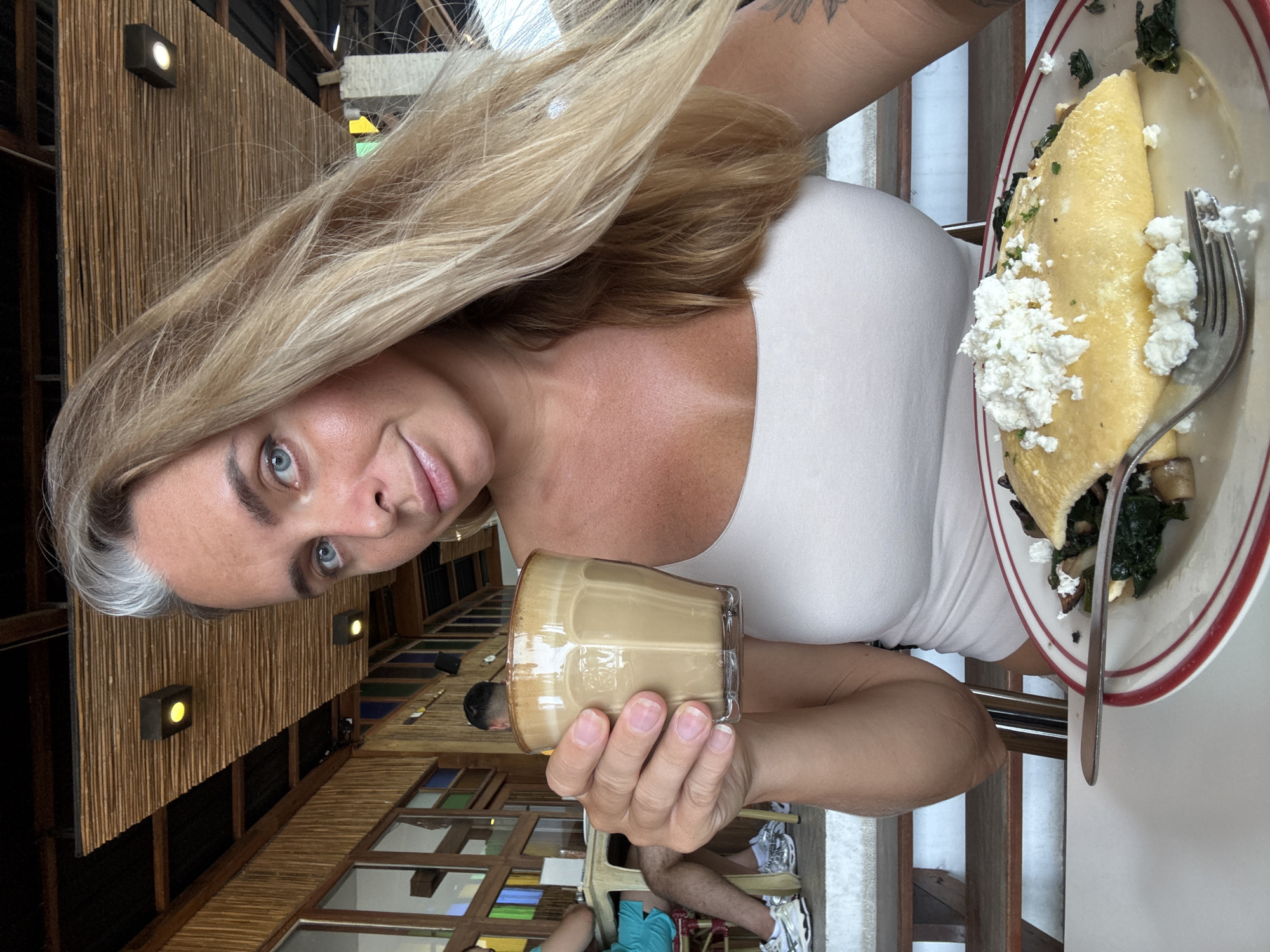 A woman with blonde hair and blue eyes is holding a glass of coffee in her left hand. She is wearing a white tank top. In front of her on a plate is an omelet topped with feta cheese and served with spinach and mushrooms. She appears to be at an outdoor cafe, with a bamboo ceiling and wooden beams visible in the background. There are other people seated at tables in the background. The overall setting suggests a tropical location.