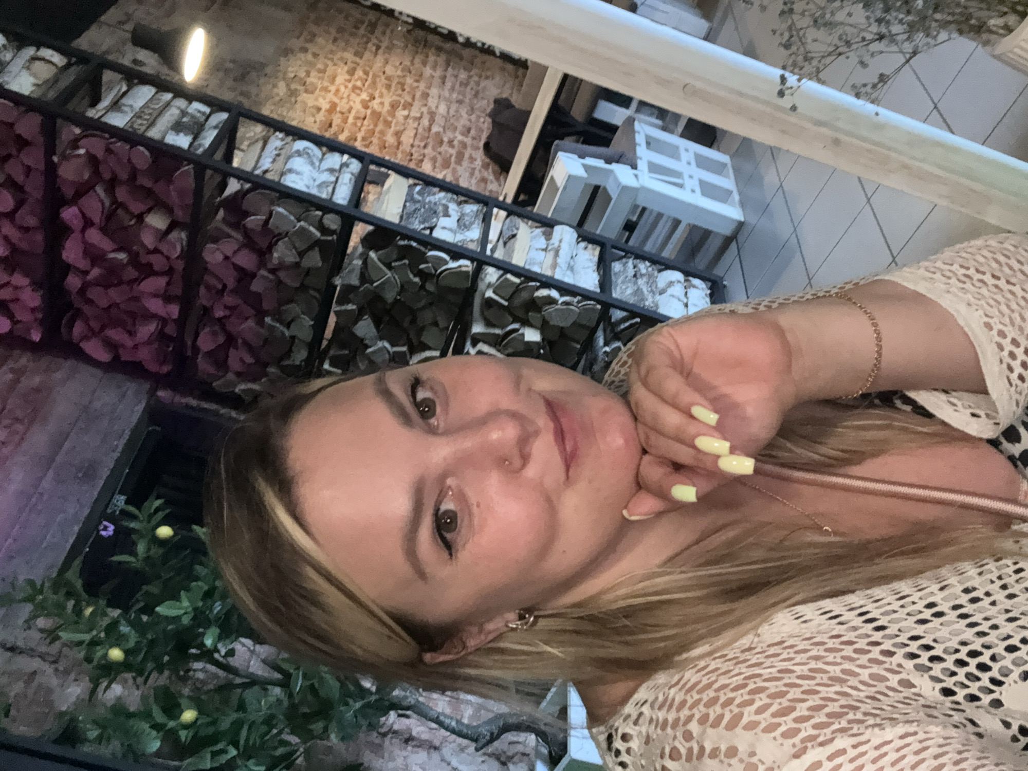 This media file is a close-up, selfie-style portrait of a blonde woman. She is leaning her head on her hand, which has bright yellow nail polish. She is wearing a white, crocheted top. Her hair is long and blonde. She appears to be indoors, with a background that includes stacks of firewood behind a black metal grate. There are also some decorative white shelves in the background, and a small lemon tree with yellow fruit. The lighting suggests it could be evening or indoors. The overall mood appears relaxed and casual. No visible text is present in the image.