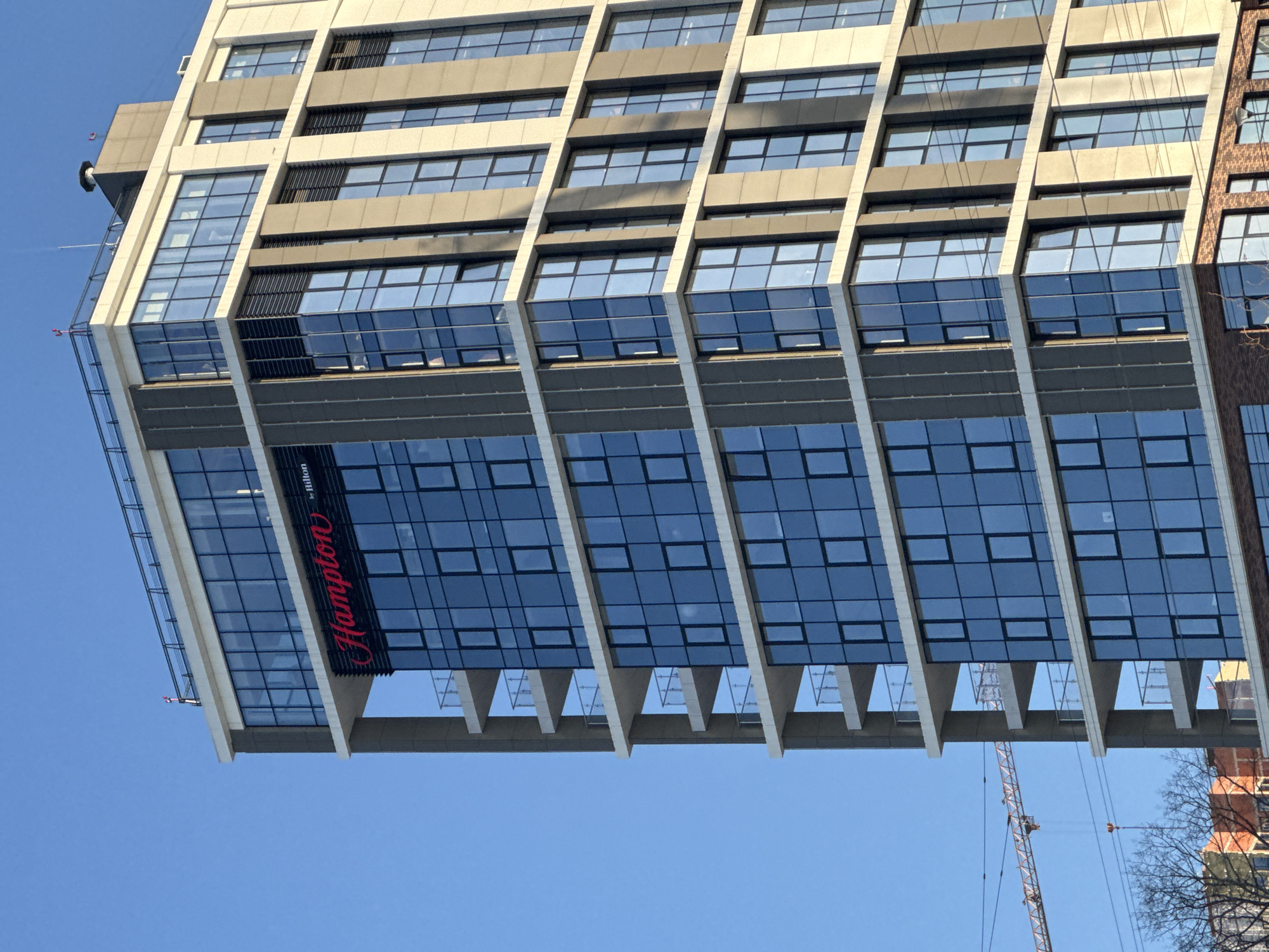 A partial, upward-angled view depicts the Hampton by Hilton Perm hotel, located at Monastyrskaya Street, 43, Perm, Perm Krai, Russia. The multi-story building features a modern architectural style, characterized by a dominant blue-tinted reflective glass facade and interspersed vertical and horizontal sections of light grey and dark grey cladding panels. The brand name "Hampton" in red script, followed by "by Hilton" in smaller white text, is prominently displayed on an upper section of the building's face. Multiple balconies with glass balustrades are visible along the left side of the structure. The building is presented under a clear, bright blue sky. In the mid-ground, to the left of the hotel, a section of an orange-colored construction crane is visible. The lower-left portion of the image shows bare tree branches and the upper segments of other, less modern buildings in the distance, including structures with visible brickwork. No individuals or specific actions are discernible within the frame.