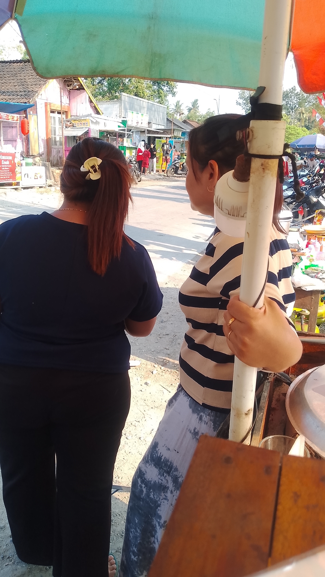 In Karanglangu, Indonesia, two women stand on a dirt road under a colorful awning. The woman on the left, viewed from behind, wears a dark blue t-shirt and black pants, her auburn hair pulled back with a white clip. The woman on the right, also viewed from behind, wears a striped long-sleeved shirt and a patterned skirt, her hand resting on a white pole. In the background, a street market bustles with activity, featuring stalls, motorcycles, and other pedestrians. The sun casts long shadows, indicating it is likely late afternoon.