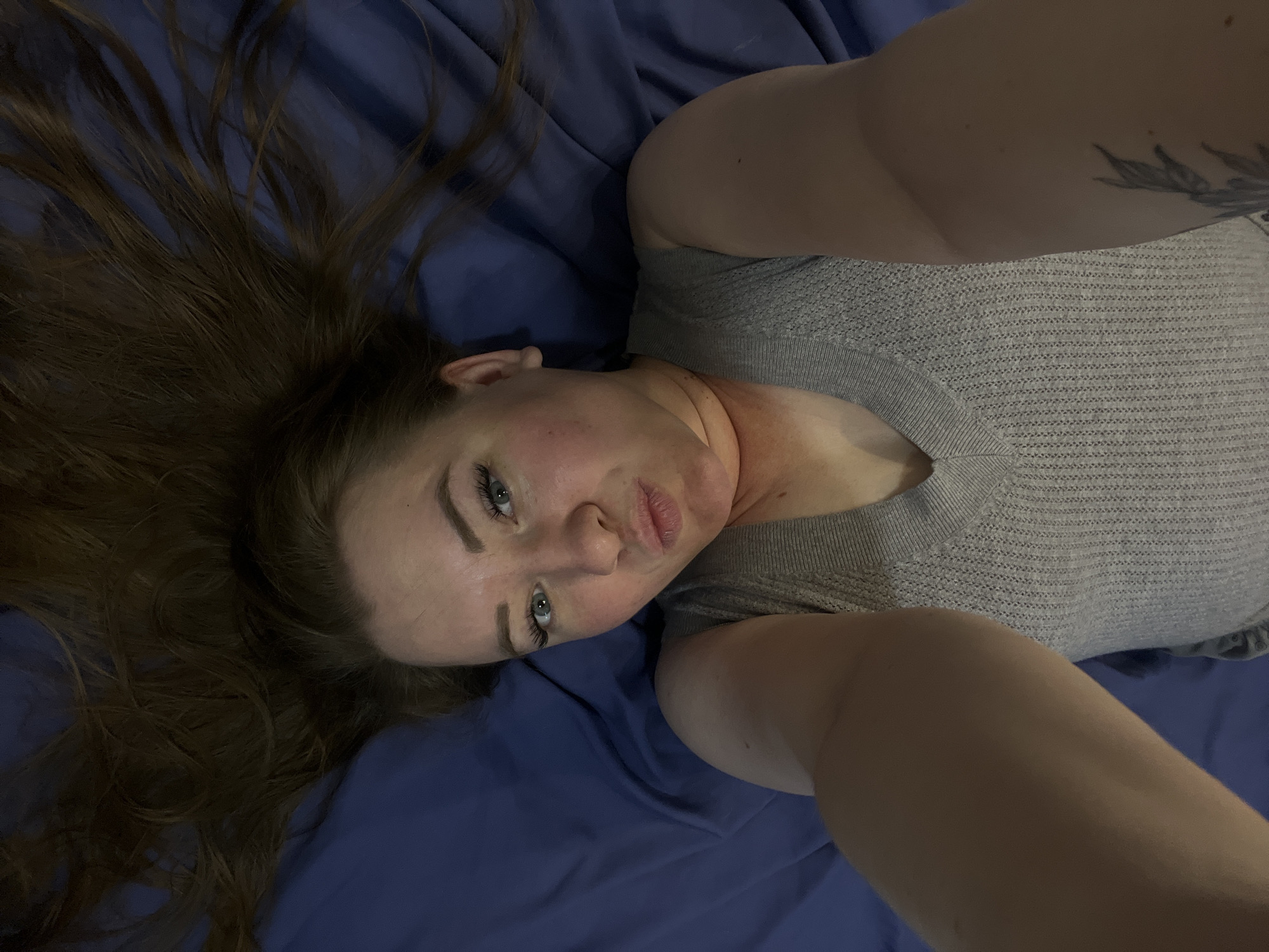 A fair-skinned woman with auburn hair is lying on a blue sheet or blanket, her head toward the left of the frame. She is wearing a gray knit top. The photograph appears to be a selfie, with the camera positioned above her. There is a tattoo visible on her right upper arm. The location, Chaikovsky, Russia, cannot be verified from the image.