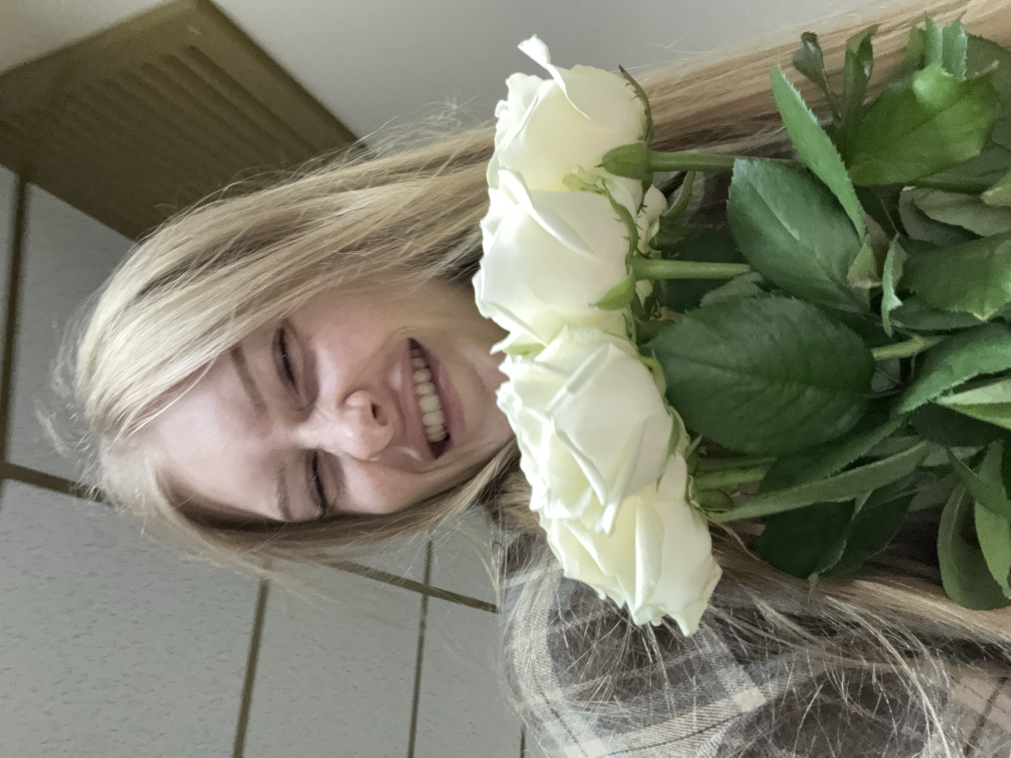 A young woman with blonde hair is smiling and holding a bouquet of white roses. She is wearing a plaid shirt. The background appears to be an indoor setting with a textured ceiling. The image is a close-up shot, focusing on the woman and the flowers. There is no discernible information to determine the location in the city of Perm, Russia.