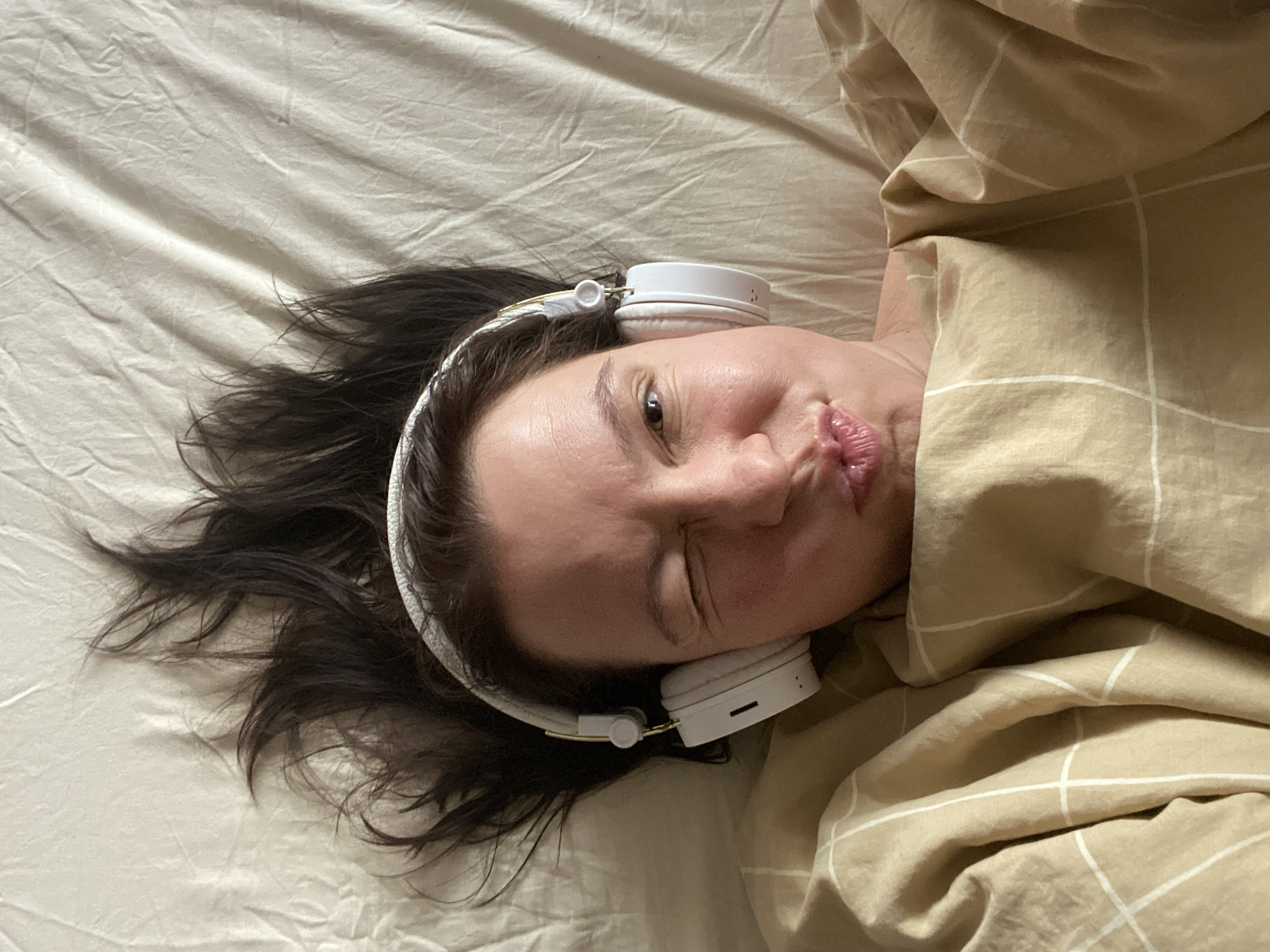 A woman with dark hair is depicted lying on her back in a bed, viewed from an overhead perspective. Her head is positioned on a light beige or off-white sheet, with her dark hair fanned out around her. She is wearing white over-ear headphones, with one earcup visible on the right side of her head, showing a small gold-colored detail and possibly control buttons.
Her face is turned towards the camera, displaying a playful expression. One eye is open and looking directly at the viewer, while the other is squinted. Her lips are distinctly puckered into a "kissing" or "duck face" gesture. The upper part of her body is partially covered by a light brown or tan duvet featuring a subtle white grid pattern. The duvet is ruffled, suggesting a relaxed, informal setting.
The overall scene suggests a moment of leisure and relaxation, possibly listening to music or an audiobook. There is no discernible text visible on the headphones or bedding. The location context provided is Chaikovsky, Russia. No specific time of day or weather conditions are indicated within the image.