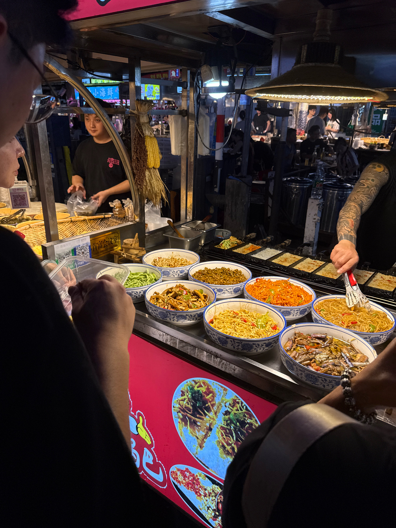 A street food vendor in Yiwu, China, is preparing food for a customer. The vendor, a young man wearing a black t-shirt with a circular logo, stands behind a counter filled with various dishes in blue and white patterned bowls. He is holding a clear plastic bag and appears to be serving a customer. In front of the counter, a person is visible from the shoulders up, wearing glasses and a black shirt, reaching for a clear plastic container. Another person's tattooed arm is visible on the right, using tongs to select food from a bowl. The background shows other stalls and people, suggesting a bustling market or food court. The scene is illuminated by overhead lights and neon signs.