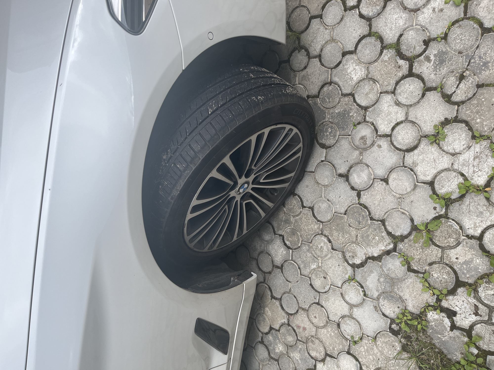 The image shows the front passenger side wheel and fender of a white BMW sedan parked on a decorative circular stone paver surface. The car's tire is partially visible, with the BMW logo on the wheel. The ground is composed of interlocking gray pavers with some sparse vegetation growing in the gaps. There is no indication of the specific location being in Ореховка, Russia; this is solely a description of the visual content of the image.