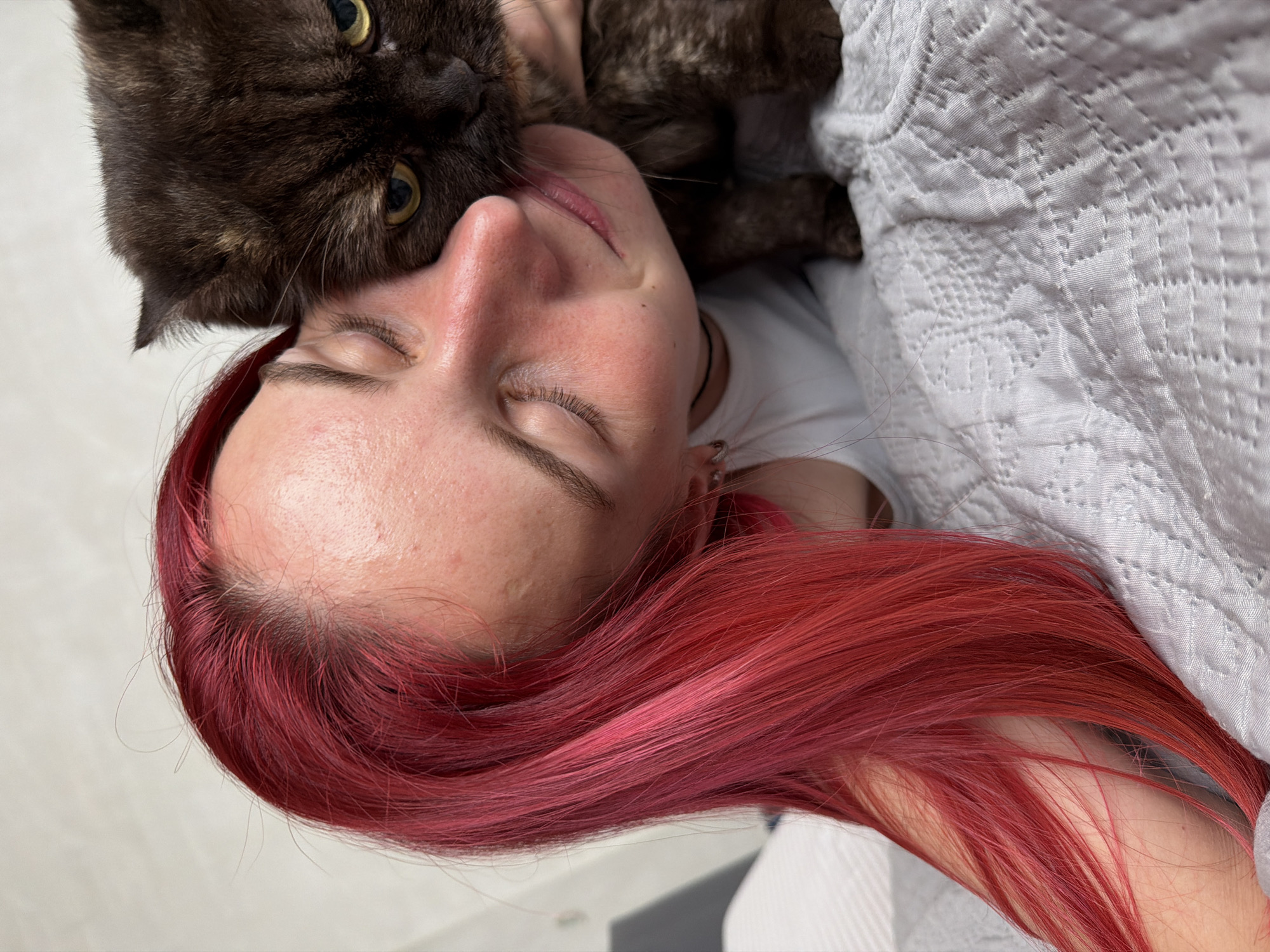 In a close-up, indoor shot, a person with vibrant red hair is affectionately nuzzling a dark brown cat. The person's eyes are closed, and they are leaning their cheek against the cat's head. The cat, with yellow eyes, appears to be looking directly at the camera with a slightly displeased expression. Both subjects are positioned on a white quilted surface, likely a bed or sofa. The background is a plain, light-colored wall. The image provides no discernible information about the city of Chaikovsky, Russia.