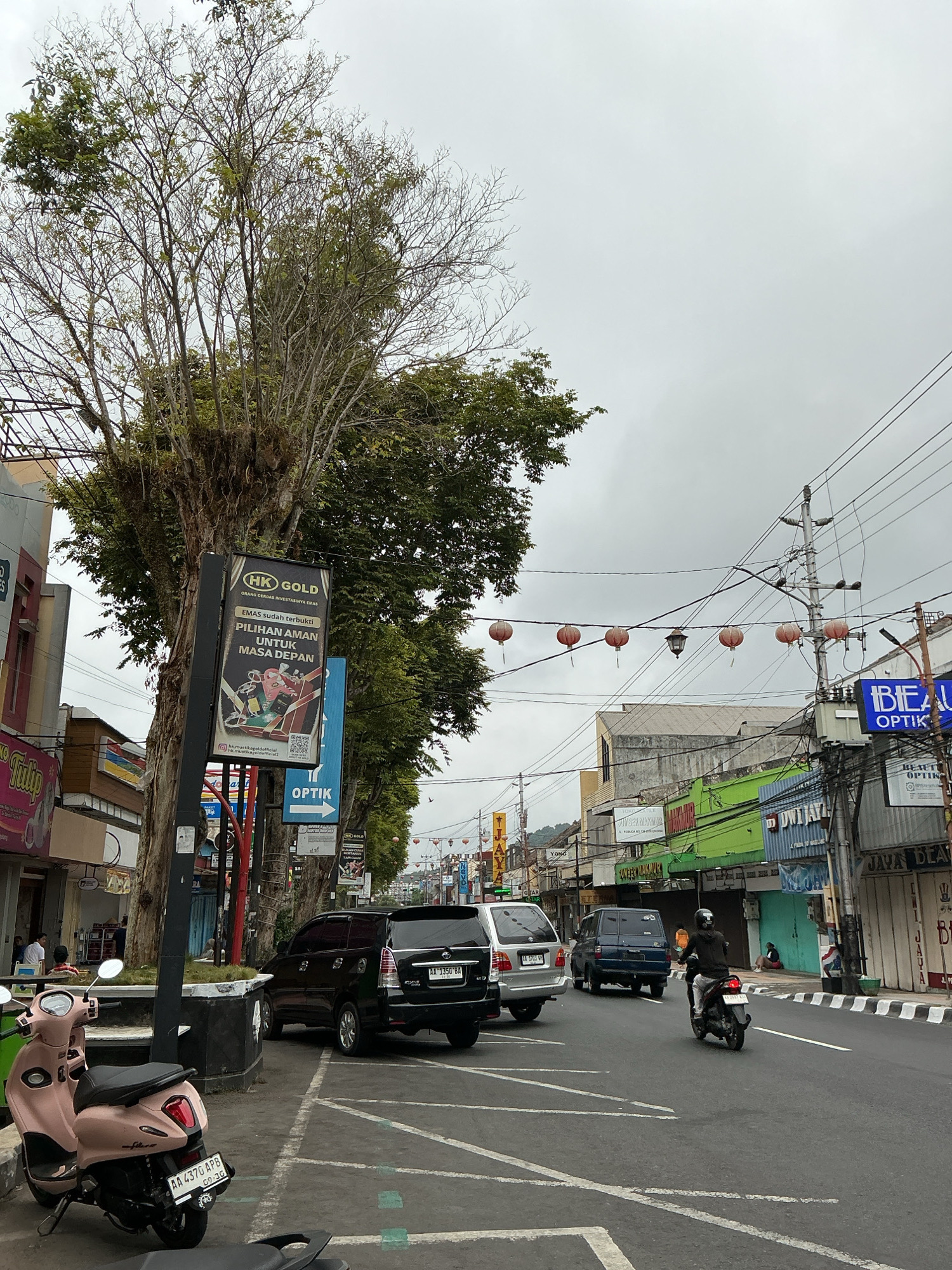 This image captures a street scene in the city of Magelang, Indonesia, under an overcast sky. The foreground features a light pink scooter parked on the side of the road, with its license plate reading "AA 4370 APB". Behind it, a black SUV with license plate "AA 1350 BA" is parked. Further down the street, a blue minivan is visible, followed by a motorcyclist wearing a black jacket and helmet, traveling away from the camera.
The street is lined with various shops, some of which have brightly colored awnings, including green and blue. One shop displays a sign for "BEAUTY OPTIK". A large tree with sparse foliage stands prominently on the left side of the street, with a black billboard attached to it advertising "HK GOLD". Red lanterns are strung across the street, adding a festive touch. Power lines and utility poles are visible throughout the scene. In the background, buildings with signs suggest a commercial area, and a person can be seen sitting on the curb in front of a shop. The road is paved and marked with white lines.