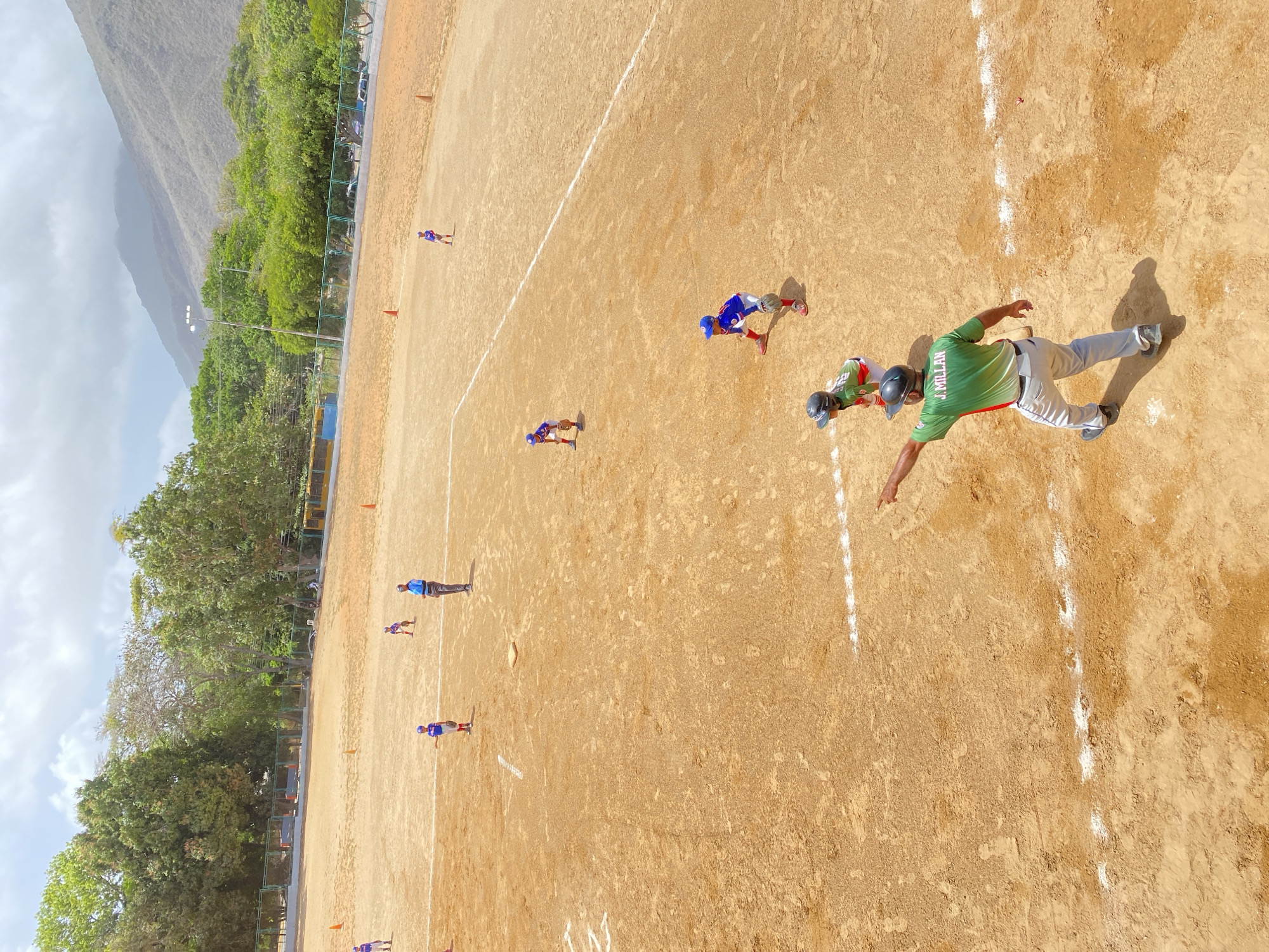 The image shows a baseball field with a group of people, likely players and coaches, on it. The field is dirt, with white lines marking boundaries. Players are wearing uniforms, some blue and some green. In the background, there are trees and hills. The location is La Asuncion, Venezuela.