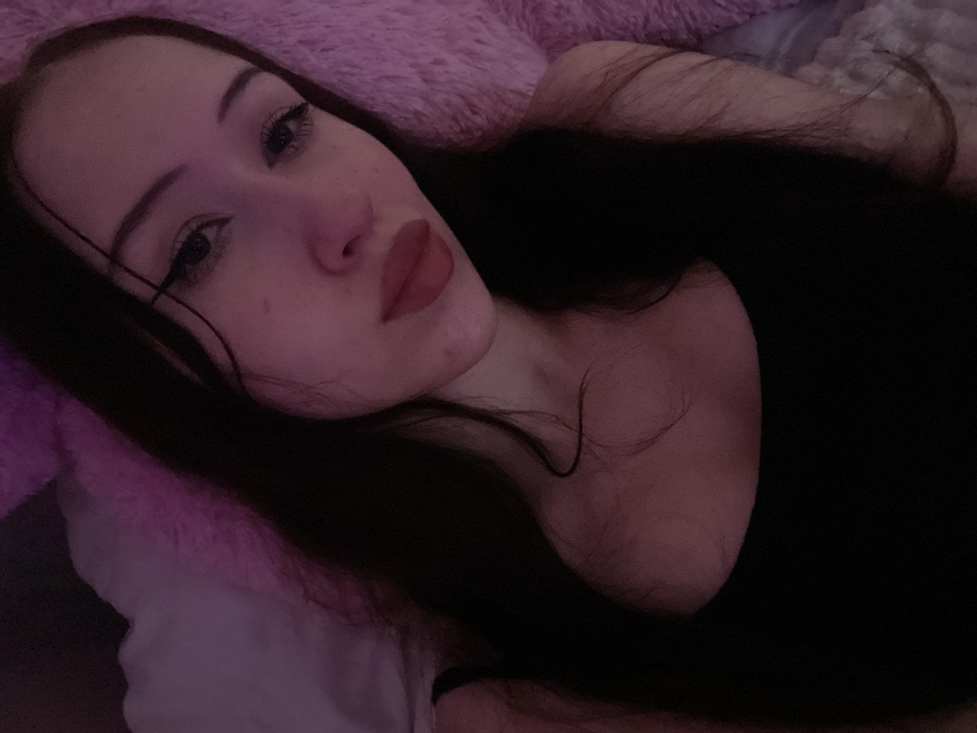 A young woman with dark hair and light skin is lying down and looking into the camera. She is wearing a black tank top and her face is partially illuminated by soft pink light. Her makeup includes winged eyeliner and lipstick. The background appears to be a fluffy pink surface. The image was taken indoors.