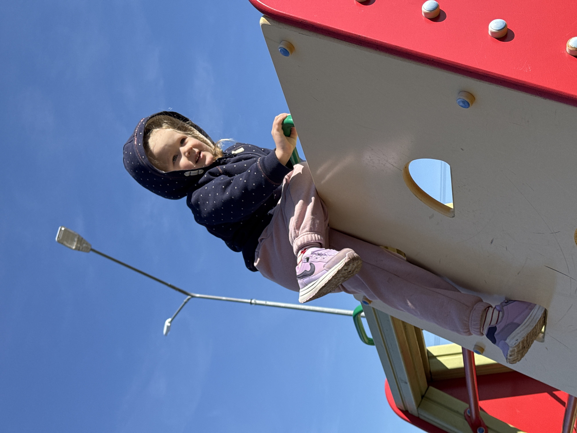 A child wearing a navy blue, polka-dot hooded jacket and light pink pants is shown hanging off a red and white playground structure. The child has blond hair peeking out from under the hood and is smiling at the camera. The child's feet are clad in lavender-colored sneakers with white soles. The child is holding onto a green handhold. The playground structure is red on top, with white sides and a circular cutout. The background consists of a bright blue sky and a street lamp. The image was taken in Горный, Russia.