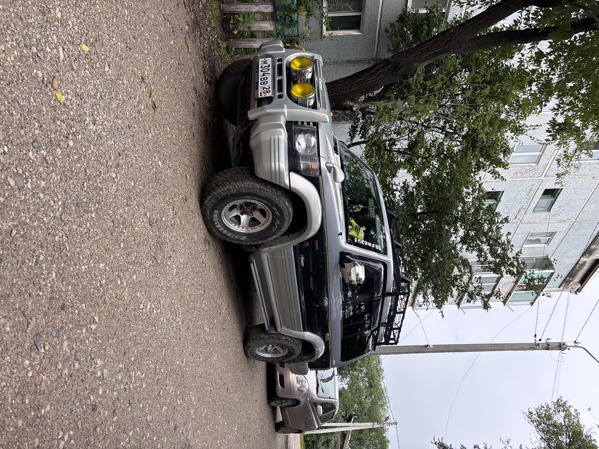 A matte gray and black Mitsubishi Pajero SUV is parked on a gravel road next to a light blue apartment building in Blagoveshchensk, Russia. The SUV has yellow fog lights on the front bumper and a roof rack. A light pink hatchback is parked behind the SUV. The apartment building has several windows and balconies. A tree with green leaves is in front of the building. The sky is overcast. The image was taken on a cloudy day.