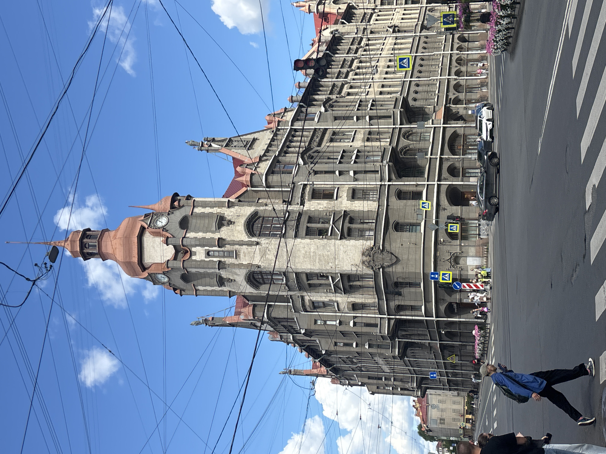 The image shows a wide street intersection in Saint Petersburg, Russia, dominated by a large, ornate building with a distinctive tower and red roof. The building exhibits a mix of architectural styles, with stone facade, arched windows, and decorative elements. Several street signs, including pedestrian crossing signs and traffic lights, are visible. A few cars are driving on the road, and pedestrians are crossing the street. The sky is blue with scattered clouds. Overhead, a complex network of electrical wires crisscrosses the scene. The image appears to be taken on a sunny day.