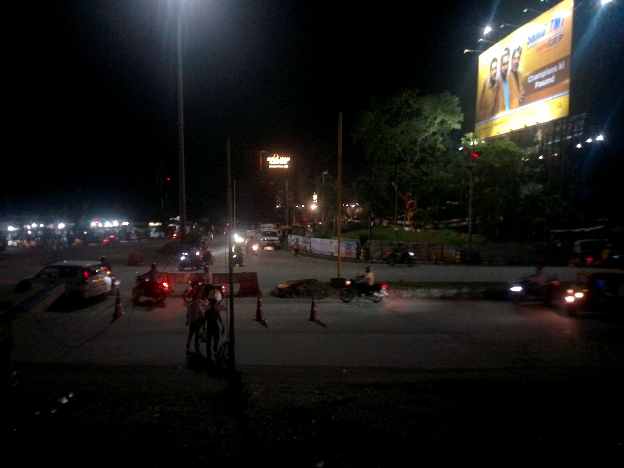 The image depicts a nighttime street scene in Siliguri, India, likely near a busy intersection. A large billboard advertising "Sang TMI Wing Grip" with images of three men is prominently displayed overhead, illuminated by bright lights. Below, traffic is visible, including cars and motorcycles. Several individuals are on foot, some walking on the side of the road and others appearing to be crossing. Two men in the foreground are standing on the road, one wearing a light-colored shirt and dark pants, the other in a dark shirt and pants. Traffic cones are placed on the road, possibly indicating construction or a diversion. The overall atmosphere is one of urban nighttime activity, with artificial lighting dominating the scene.