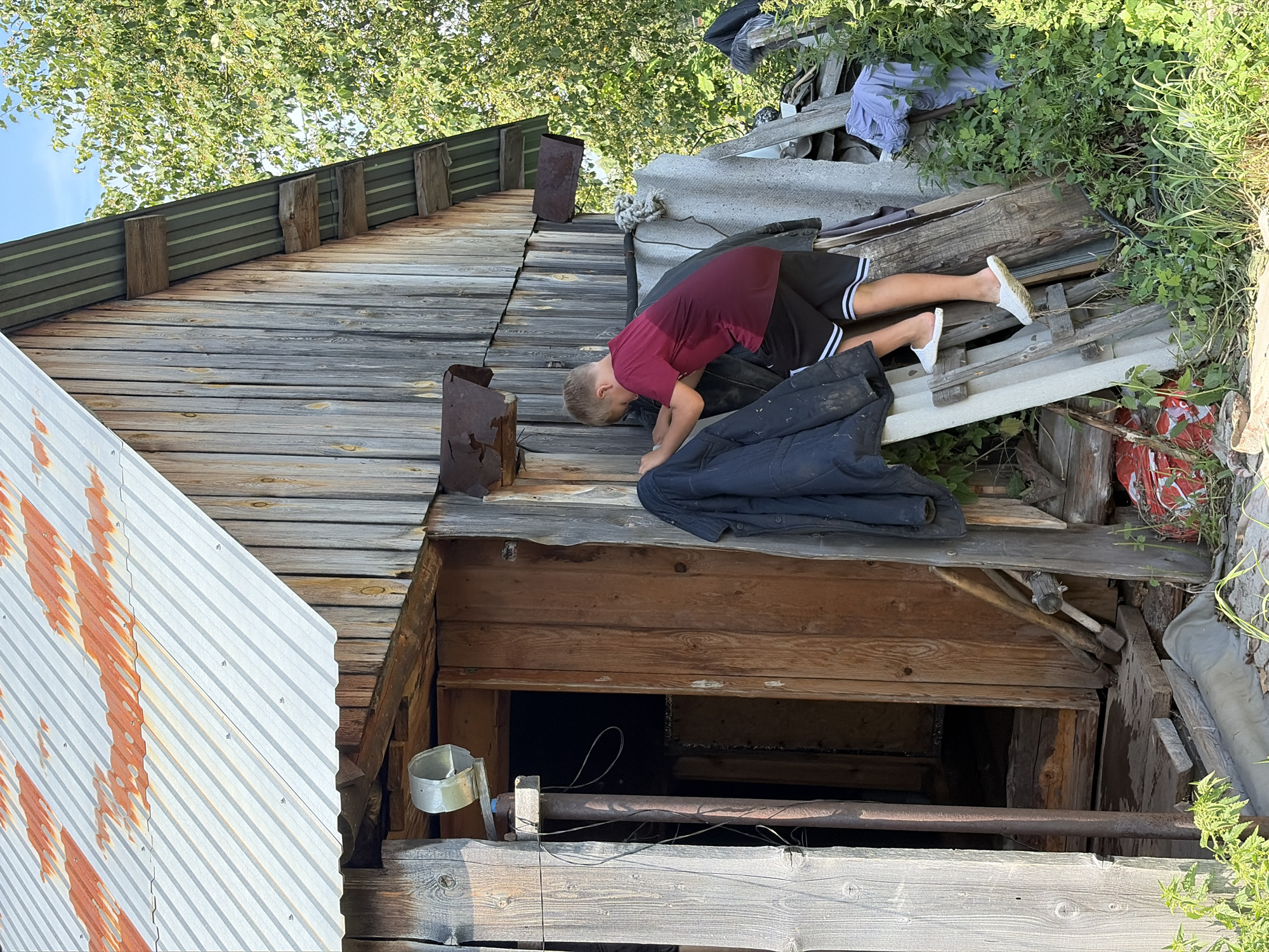 A young boy wearing a burgundy t-shirt and black shorts with white stripes is climbing up a makeshift ladder made of wood and corrugated metal sheets. He is positioned on the right side of the image, facing away from the camera and towards a rustic wooden structure that appears to be a shed or outbuilding. The structure has a corrugated metal roof with visible rust and weathered wooden walls. A dark-colored jacket is draped over the side of the structure near where the boy is climbing. The area is overgrown with green vegetation and appears to be outdoors, possibly in a rural or semi-rural setting. The ground is covered with debris, including pieces of wood and what looks like a red plastic container. The sky is visible in the upper right corner, indicating it is daytime. The location is identified as Kemul, Russia.