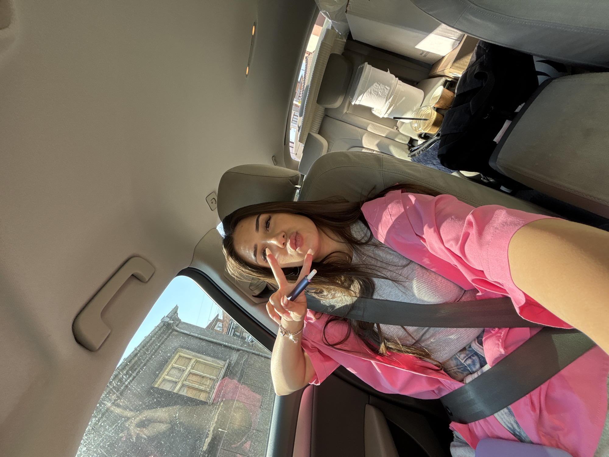 The image captures a young woman seated in the front passenger seat of a vehicle on a bright, sunny day in the city of Rakovski, Bulgaria. The woman has long, dark hair and is dressed in a pink collared jacket over a light grey ribbed top, with denim fabric visible below the jacket. She is secured by a black seatbelt across her chest and lap. Her right hand, adorned with a silver-toned bracelet on her wrist, holds a dark blue and black vape pen while simultaneously forming a two-finger peace sign. Her lips are pursed in a pouting expression, directed towards the camera, indicating a self-portrait.
The car's interior features light-colored fabric upholstery and a matching headliner. Through the window to the left of the woman, an older, two-story grey building with a weathered exterior and a prominent wooden window frame is visible under a clear blue sky. A section of a lower, adjacent building is also discernible.
The back seat of the car is loaded with various items, including multiple white cardboard boxes, stacked white fabric rolls or bedding, a black bag, and two white plastic buckets, one stacked atop the other. Two disposable coffee cups, one with a straw, are positioned on the floor or console between the front and back seats. A purple object, possibly a phone case or bottle, rests near the woman's lap.