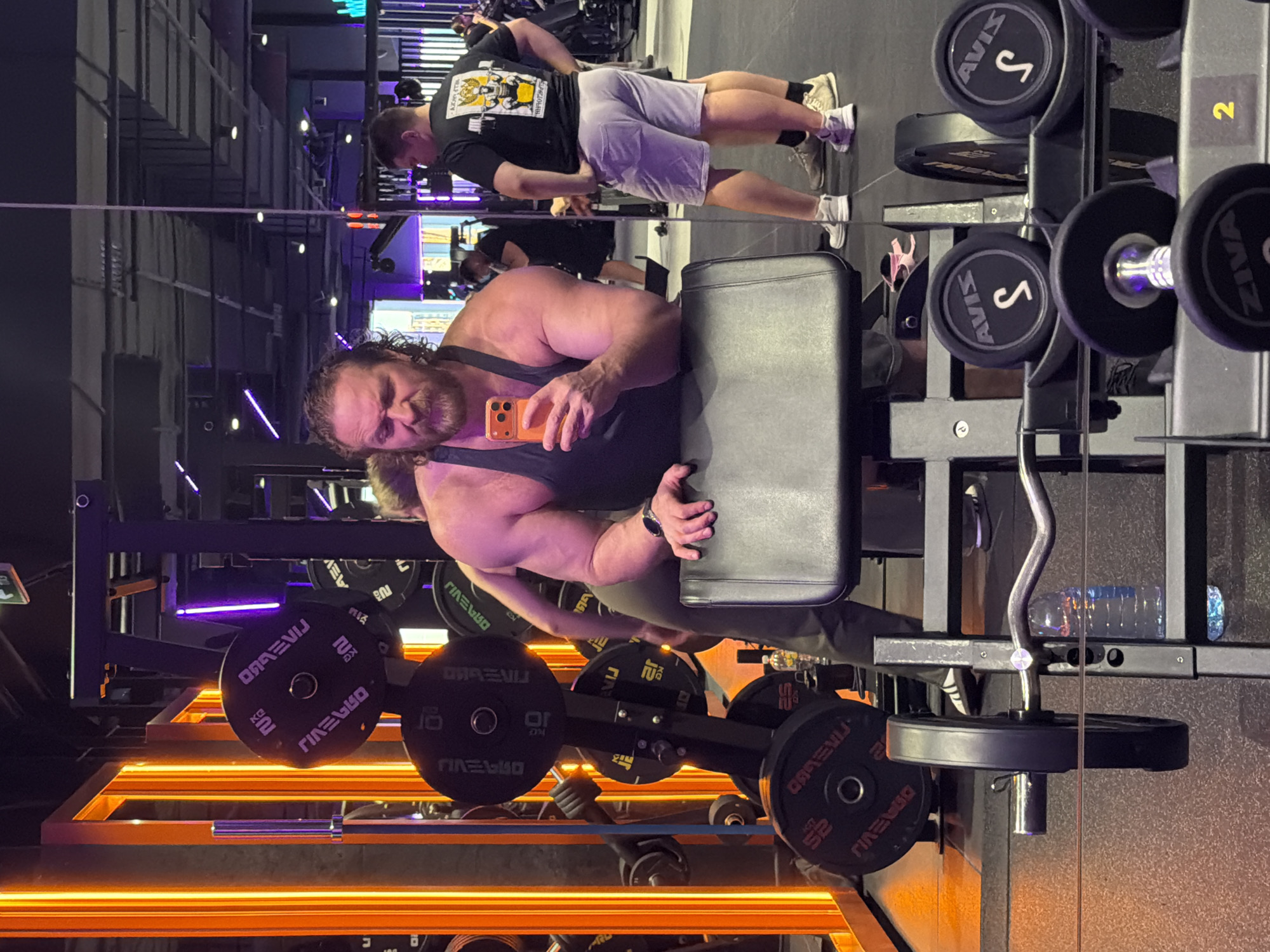 This media file captures a scene inside a modern gym in Saint Petersburg, Russia. In the foreground, a muscular man with a beard and long curly hair, wearing a dark tank top, takes a selfie in a large mirror using an orange smartphone. He has a focused expression and wears a watch on his left wrist.
Reflected in the mirror, further back, another man is visible. He wears a black t-shirt with a yellow graphic design and light-colored shorts, standing upright, possibly between sets or posing.
The gym features dark walls, contrasting bright orange linear lights near the floor, and subtle purple accent lighting. Various weightlifting equipment is present, including a rack of dumbbells on the right side of the frame, and barbell weight plates stored on a lower rack. An EZ curl bar and a water bottle are also visible.
Visible text includes brand names and weight indicators on the equipment. On the dumbbells, "AVIS S" and "ZIVA" are discernible, along with the number "2" on a dumbbell rack. On the weight plates, the brand "DRAEVIJ" is repeated, with weights such as "2.5 KG", "5 KG", "10 KG", and "25 KG" clearly marked (some appear mirrored due to the reflection). The man's t-shirt has a yellow graphic featuring two stylized figures and some text, but it is largely indistinct. The overall atmosphere suggests an active fitness environment.