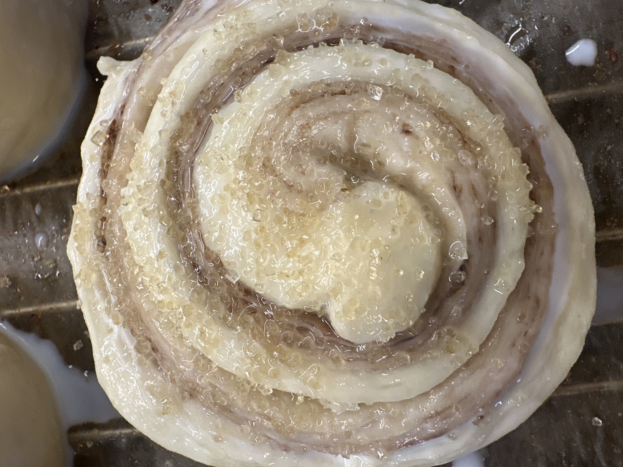 A close-up, top-down shot shows a single, large cinnamon roll on a baking sheet. The roll is swirled, with a light tan and white dough and a generous coating of large sugar crystals. There are visible layers and a slight glaze on the dough. A small amount of white icing is visible near the roll, suggesting others were iced nearby. The location, Bac Nha Trang Ward, Vietnam, is not visually supported by the image itself.