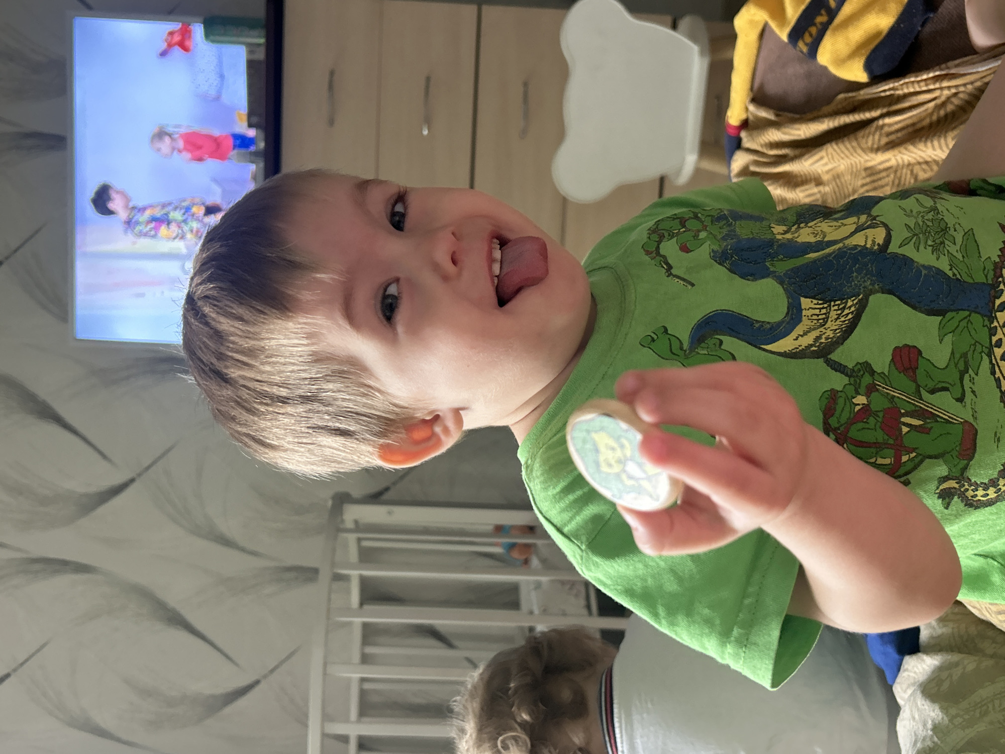 A young child is seen in a medium shot, wearing a green Teenage Mutant Ninja Turtles t-shirt, holding a small, round object. The child is sticking their tongue out. In the background, there is a television displaying animated figures, a wooden dresser, a white chair, a baby crib, and another individual in a white shirt. The photograph was taken indoors in Chaikovsky, Russia.