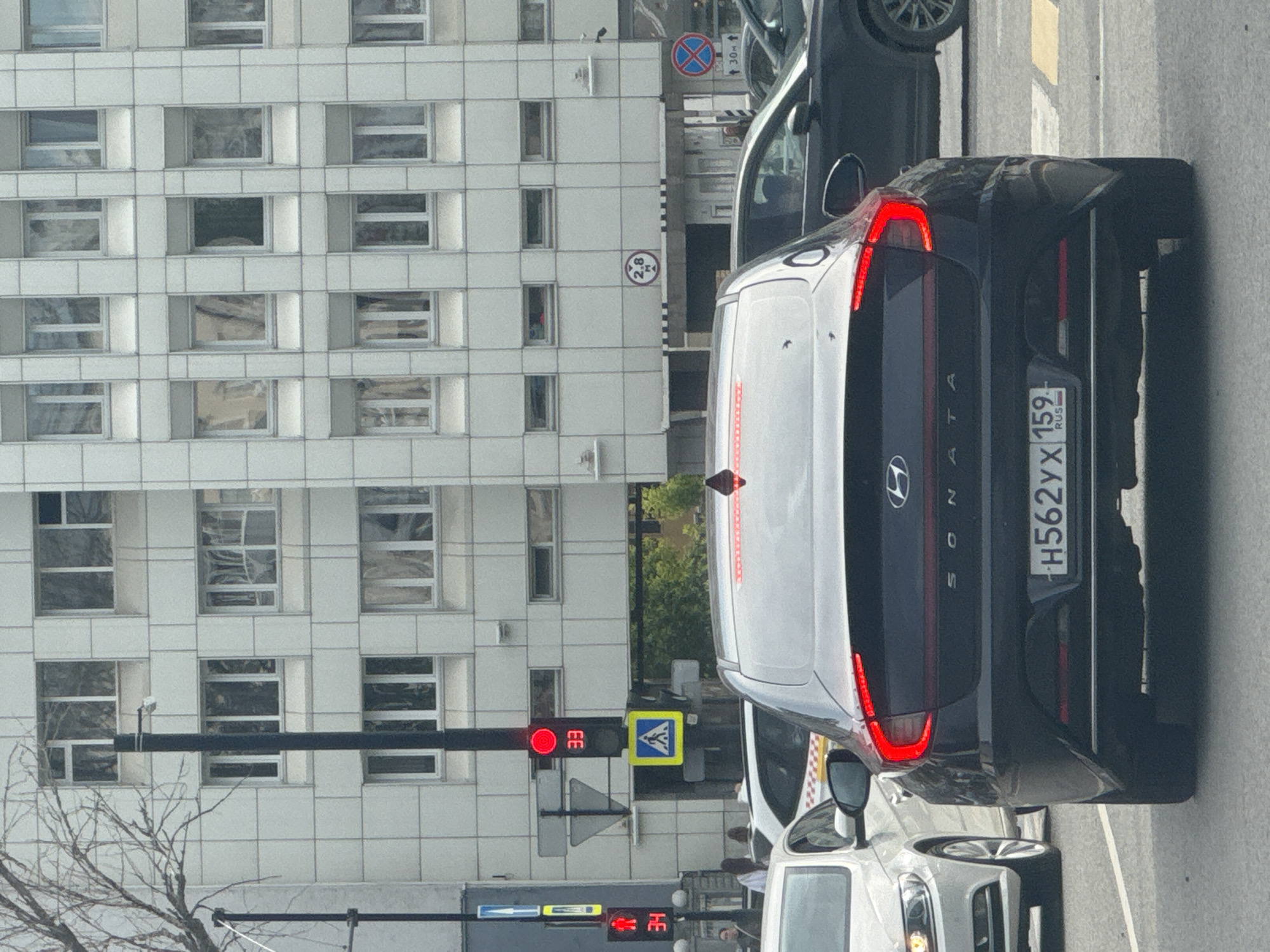 The image shows a street scene in Perm, Russia, focusing on the rear of a black Hyundai Sonata. The car is stopped at a traffic light, indicated by the red light displaying "33" seconds remaining. A pedestrian crossing signal shows a red walking figure with "34" seconds. Behind the Sonata, other vehicles are visible, including a white car on the left and a darker car on the right. The background features a multi-story building with a grid-like facade of windows. Traffic signs, including a height restriction sign of "2.8 m" and a "no entry" sign, are present. The overall environment suggests an urban setting with vehicular traffic.