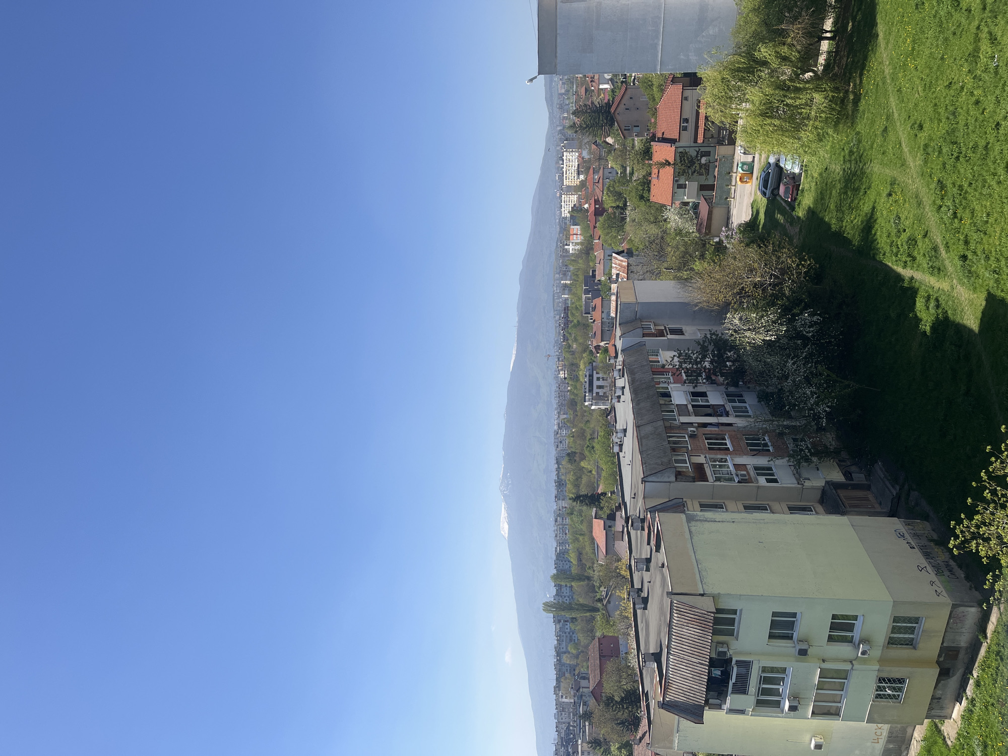 An aerial view captures a portion of Sofia, Bulgaria. Buildings with varied architectural styles and roof types dominate the foreground, giving way to a sprawling landscape with trees and residential areas. A mountain range, partially covered in snow, stands in the background. The sky is predominantly clear and blue. The scene seems to be shot during daylight hours under favorable weather conditions.