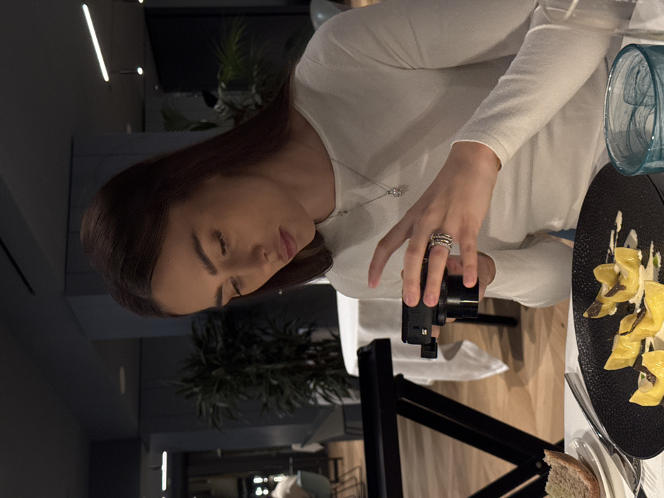 The image shows a female individual seated at a table in a modern restaurant located in Torbole, Italy. The subject, possessing long brown hair and wearing a white long-sleeved top with a silver necklace and multiple rings on her left hand, is actively holding a black compact digital camera with both hands, aiming it downwards. Positioned directly in front of her is a black textured plate containing yellow pasta shapes garnished with a white sauce and dark shavings, presumably truffles. To the left, a slice of bread rests on a small white plate, and a clear glass with a distinct teal swirl pattern is visible on the right. The table is covered with a white tablecloth. The background features a restaurant interior characterized by dark walls, a light wooden floor, potted green plants, additional dining tables with white coverings, and modern lighting fixtures, including recessed and strip lights. Other patrons are faintly discernible in the blurred distance.