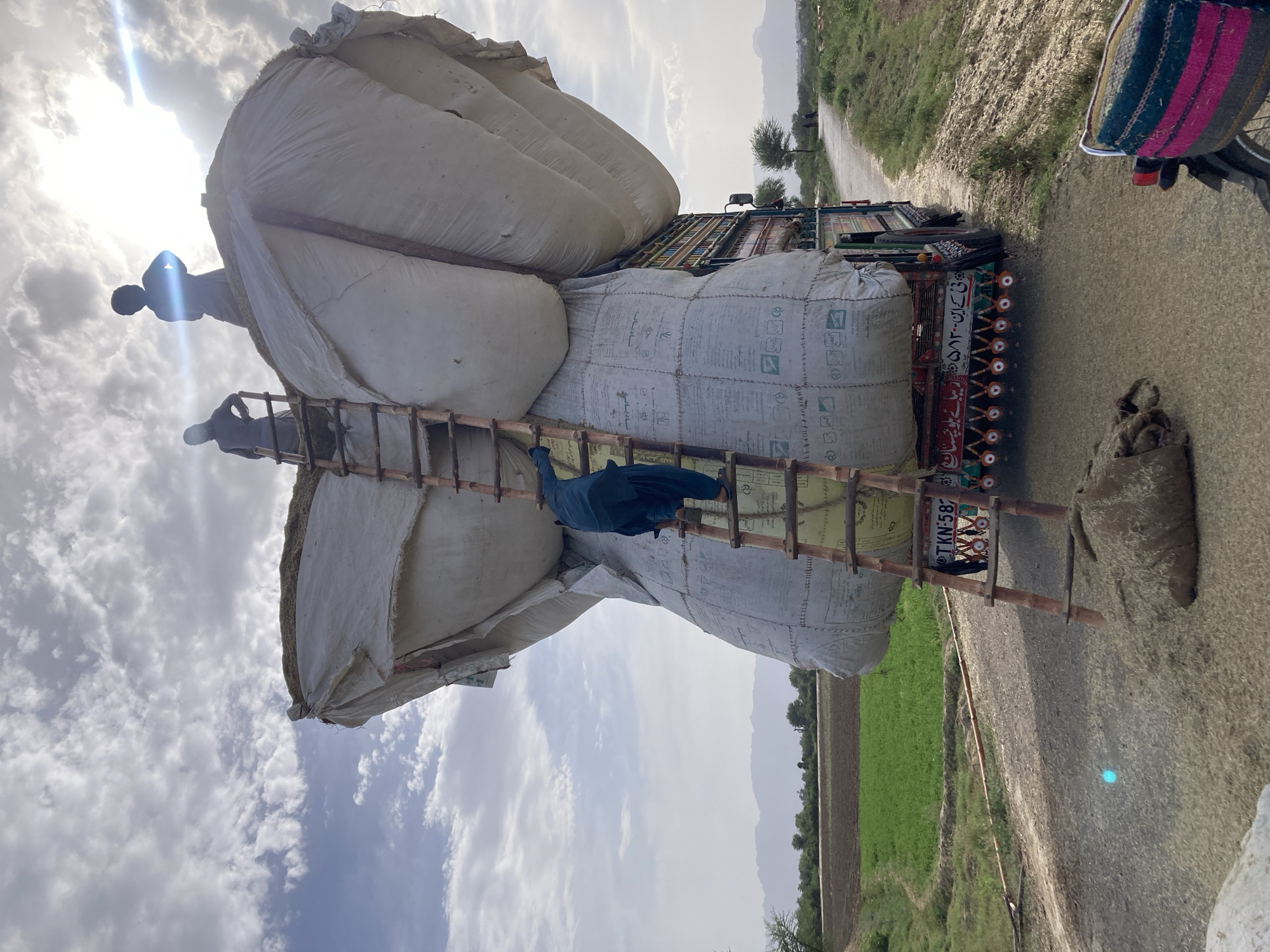 In Rāwāni, Pakistan, a truck is heavily laden with large white sacks, stacked high and secured with rope. A man in dark clothing stands on top of the sacks, while another man stands on a wooden ladder leaning against the load. A third person is positioned on the ladder, partially obscured by a blue garment. The truck is parked on an asphalt road next to an agricultural field. The sky is partly cloudy with a bright sun.