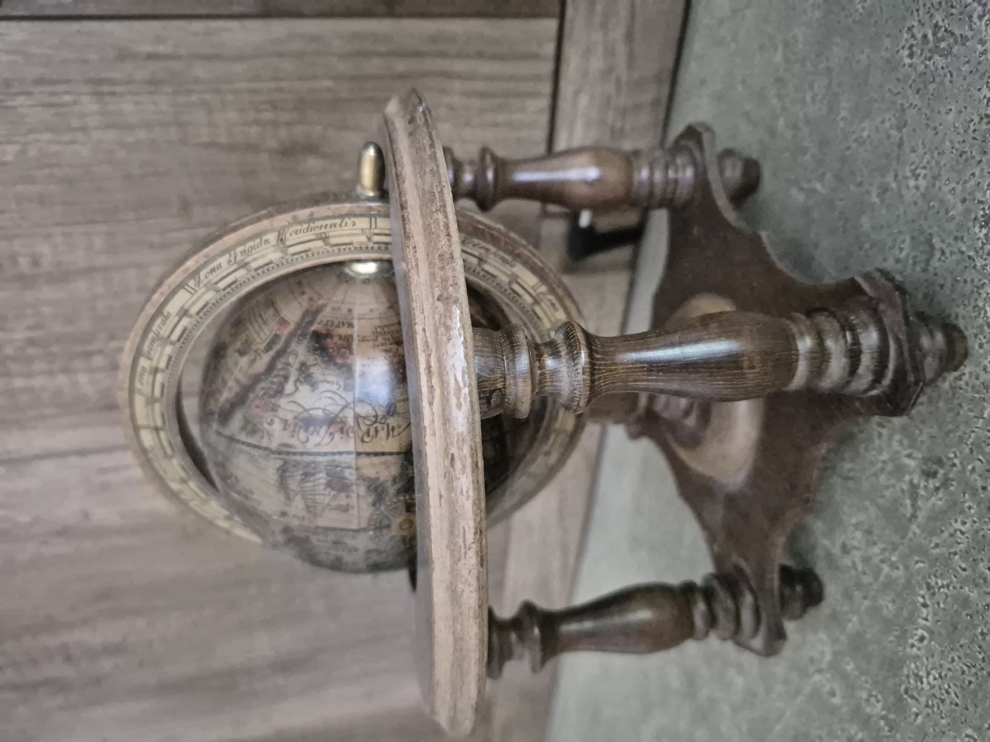 A close-up, eye-level shot depicts a small, antique globe with a wooden stand. The globe itself is spherical and features intricate, faded illustrations of landmasses, seas, and decorative elements. A brass meridian ring encircles the globe. The globe rests on a circular wooden platform supported by four turned wooden legs, which are attached to a cross-shaped base. The wood of the stand is dark and appears weathered. The background consists of a vertically oriented wooden surface, possibly a cabinet or wall panel, and the floor is a textured, speckled gray surface. The lighting is soft, casting subtle shadows. The image does not provide any specific geographical markers or context to indicate a location in Trier, Germany. There are no people present in the image.