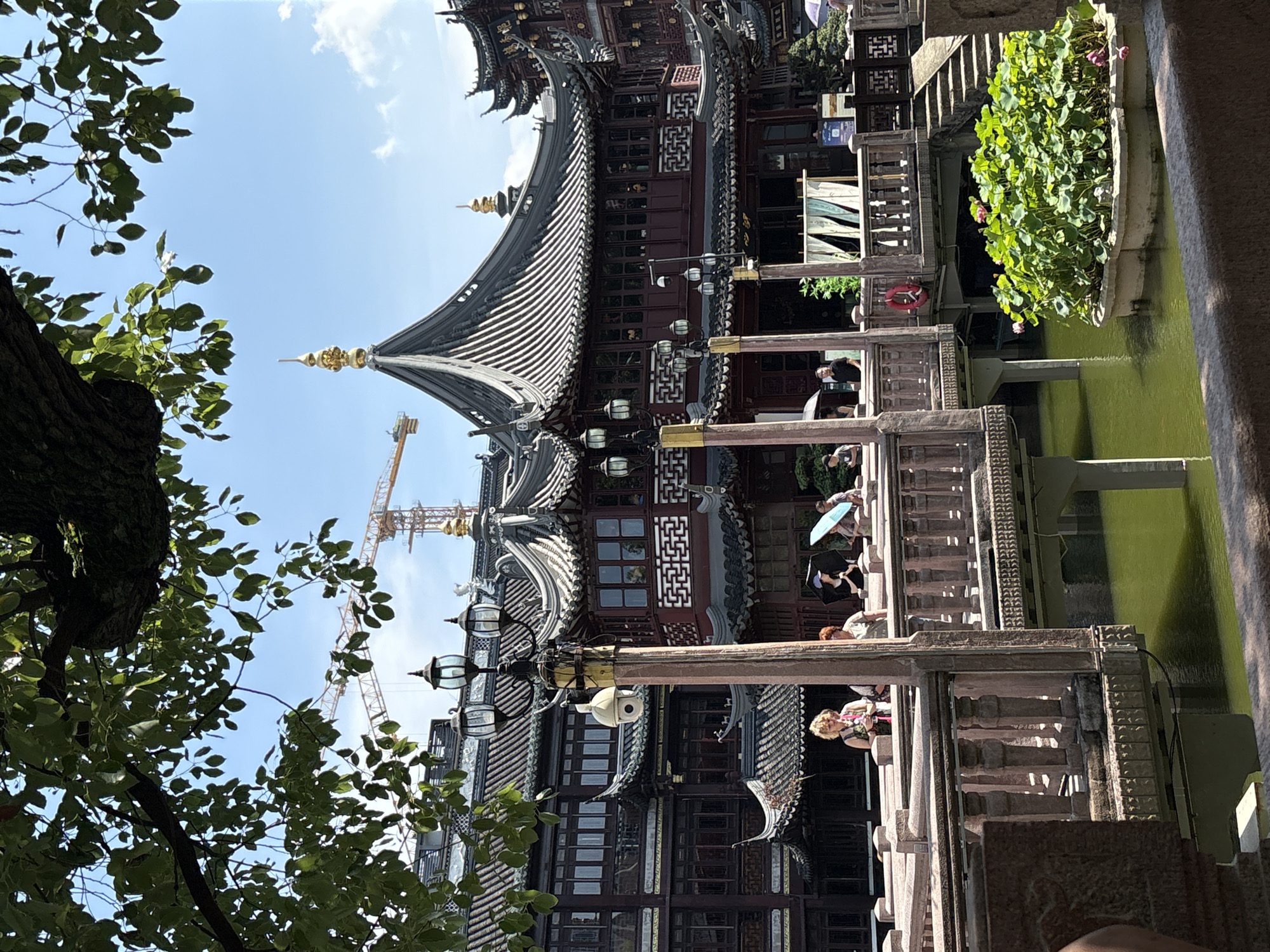 The image shows a sunny day in Shanghai's Yu Garden, a historic Chinese garden. The foreground features a stone railing overlooking a tranquil green pond filled with lotus plants. A wooden bridge, adorned with ornate lanterns and lampposts, spans the water, connecting to traditional Chinese buildings with dark tiled roofs and red accents. Several people are visible on the bridge, some holding umbrellas against the sun. In the background, a construction crane is visible against the blue sky, suggesting ongoing development in the city. The architecture is a blend of historical Chinese design and modern elements, characteristic of Shanghai's dynamic urban landscape.