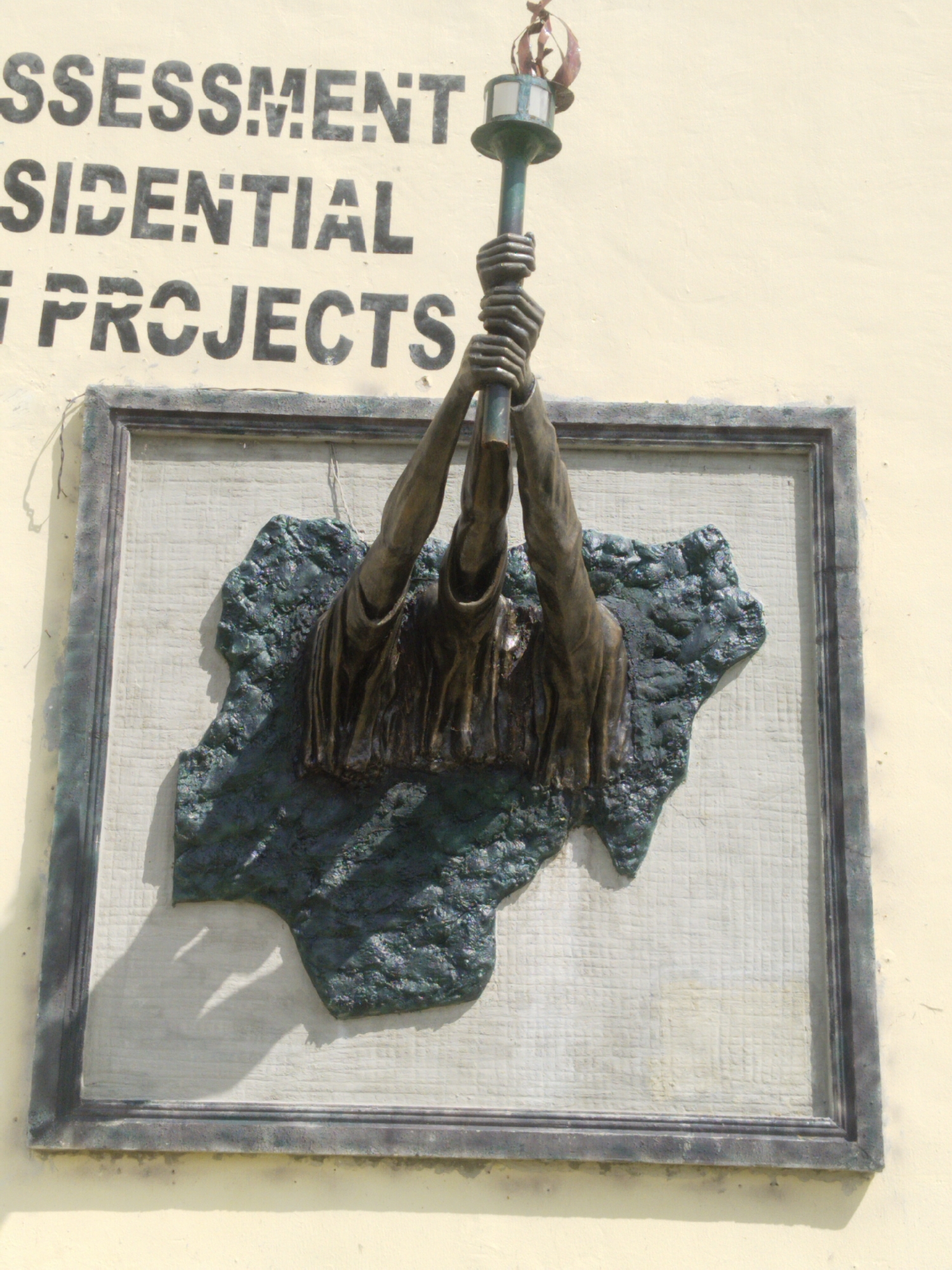 A plaque mounted on a light yellow wall shows the geographical outline of Nigeria in dark green textured material. Emerging from the center of the shape are two bronze-colored arms holding aloft a lamp or torch. The plaque is framed in a dark grey border. Above the plaque are black stenciled words, “ASSESSMENT”, “RESIDENTIAL”, and “PROJECTS”. The location is in Uyo, Nigeria.