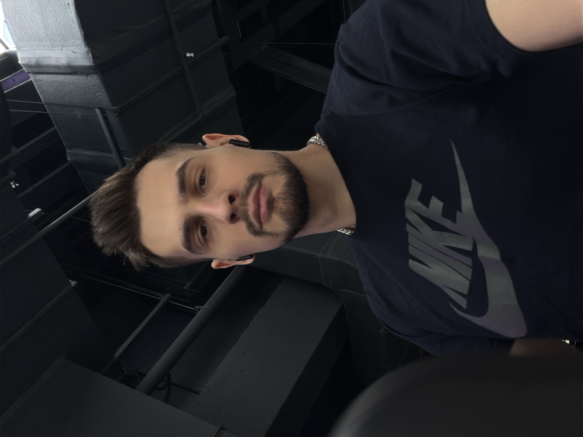 A man with dark hair and a beard is photographed indoors. He is wearing a dark-colored t-shirt with a prominent Nike logo and a silver chain. He appears to be wearing wireless earbuds. The background features a dark, industrial-looking space with various beams and structures. The photo seems to be taken from a low angle. There is no explicit information confirming the location in Krasnoyarsk, Russia.