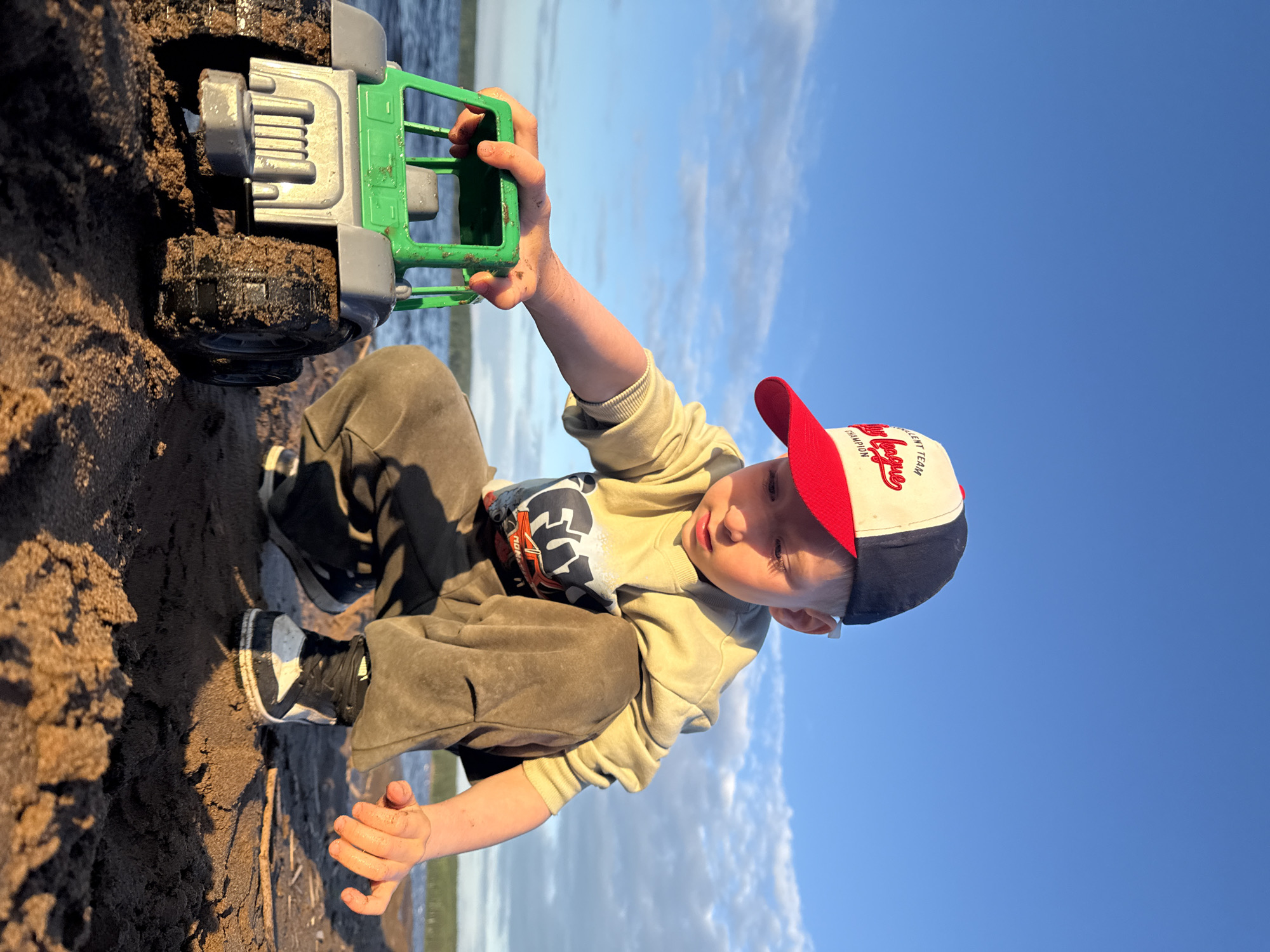 The image shows a young boy engrossed in playing with a toy truck on a sandy or muddy shoreline, likely in Chaikovsky, Russia. The boy is positioned in the foreground, crouching down, with his attention fully on the green and gray toy truck he holds. His hands and the truck's wheels are visibly covered in dirt or sand, indicating active play.
The boy is dressed casually in a light-colored long-sleeved top with a graphic design (partially visible text like "MONSTER") and dark sweatpants. He wears black and white sneakers. A red, white, and blue baseball cap adorns his head, featuring the text "Little League" and "CHAMPION" on the front. His facial expression appears focused and content.
The setting is outdoors, with a clear expanse of blue sky dotted with white clouds occupying the upper portion of the frame. A body of water, likely the Kama River or Votkinsk Reservoir, stretches across the horizon in the background. The foreground consists of dark, wet sand or mud, illuminated by warm, golden light, suggesting late afternoon or early evening. The overall atmosphere is one of peaceful, childlike engagement with nature and play.