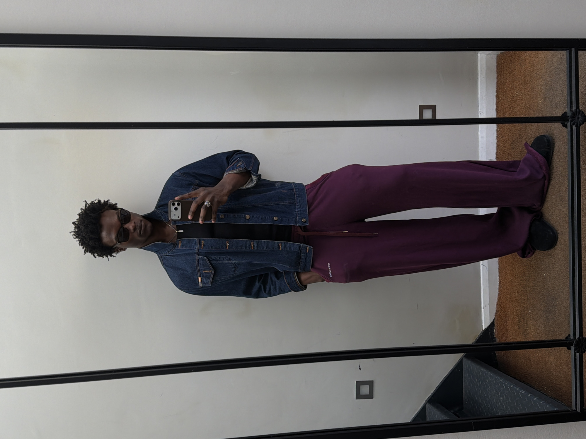 An individual is captured taking a mirror selfie indoors, likely within a residence in London, United Kingdom. The person, an adult with dark skin and short, curly dark hair, is wearing dark sunglasses that obscure their eyes. They are holding a smartphone, possibly an iPhone, in their right hand, with a distinctive ring visible on one finger.
The person is dressed in a casual yet stylish outfit. They are wearing a dark blue denim jacket over a black t-shirt. The lower body is clad in loose-fitting, wide-leg dark purple or maroon trousers. On the left thigh of the trousers, text is visible, which appears to be "IVY PARK" written in reverse, indicating a brand name. Black shoes complete the attire.
The selfie is taken in a large mirror with a prominent black frame. The reflection shows a plain, light-colored wall (off-white or beige) forming the main background. To the right of the person's legs in the reflection, a section of textured brown or tan carpet is visible, alongside a glimpse of what appears to be black stairs. A small, dark square object, possibly an electrical outlet or switch, is also visible on the wall in the reflection. The overall lighting is bright and even, characteristic of an indoor setting.