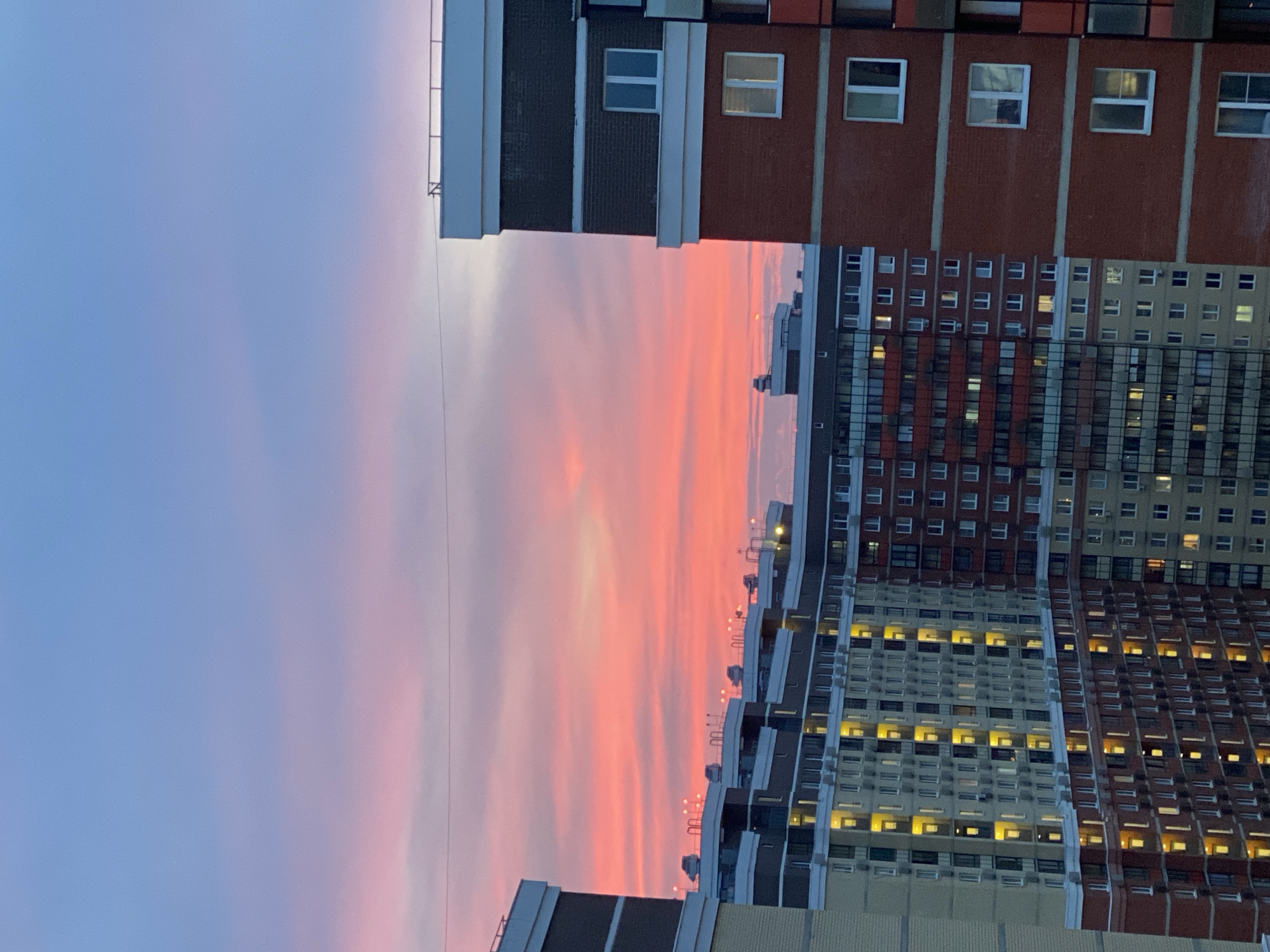 This is a vertical, eye-level, medium shot of apartment buildings against a vibrant sunset sky. The sky is a gradient of colors, transitioning from a soft blue at the top to a bright pink and orange near the horizon, with streaks of lighter pink and white clouds scattered throughout. The buildings are modern, multi-story residential structures with many windows.
The building on the right is primarily brick-red with white trim and dark accents, featuring several visible windows, some of which are lit from within. The building to the left of it is a combination of light-colored brick and dark facade sections, also with numerous lit windows. Further left, another building with a lighter facade is visible, with many windows illuminated by a warm yellow light. The rooftops of the buildings show various structural elements like vents and railings, with some cars or vehicles parked on the upper levels. The overall scene suggests an urban environment in Мирный, Russia, at dusk.