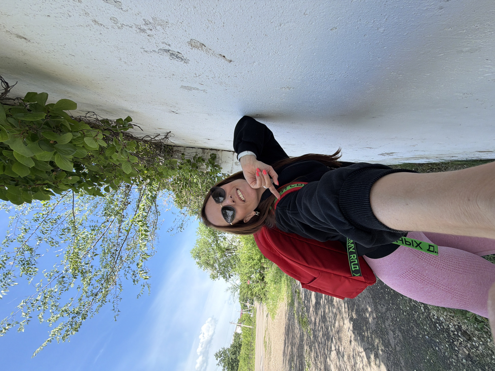 In Birobidzhan, Russia, a young woman poses for a selfie against a textured white wall. She is wearing a black long-sleeved sweatshirt, pink leggings, and sunglasses. A red backpack is slung over her left shoulder. Green ivy vines cascade down from above the wall, and a tree with green leaves is visible in the background. The sky is blue with scattered clouds. The ground is covered with gravel and patches of grass.