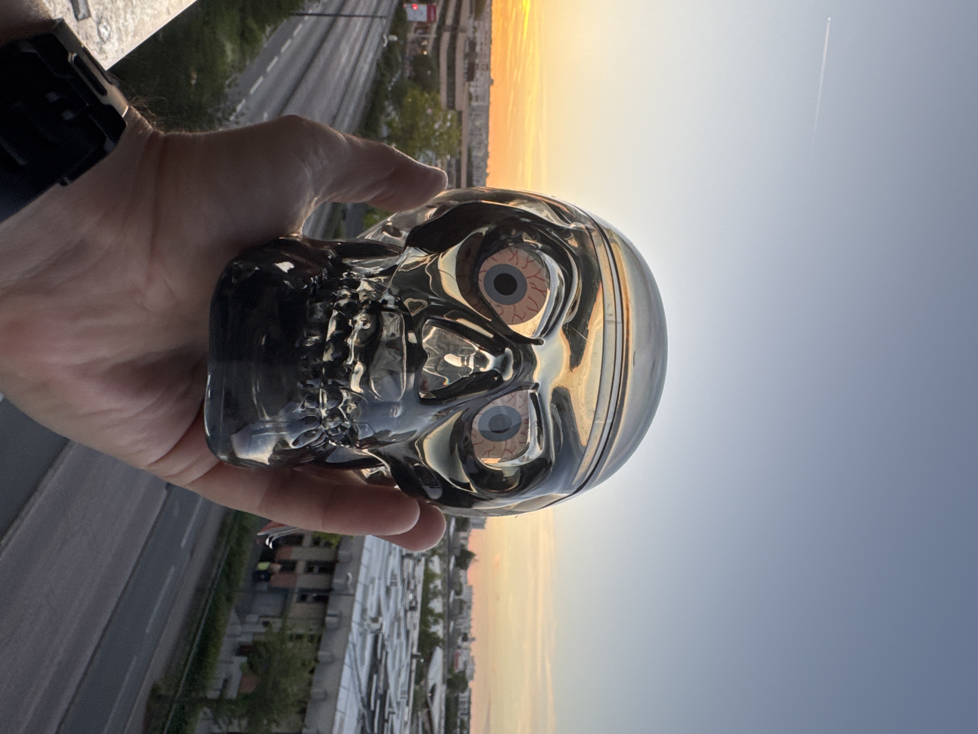 A silver, skull-shaped object with red and white eyeballs is held in a person's right hand. The object is reflective, showing the sky and buildings. The hand is positioned in the foreground, obscuring some of the skull. A black smartwatch is visible on the person's left wrist. The background consists of a city skyline at sunset, with buildings and a road. The sky is clear with a light orange glow near the horizon, and a faint contrail from an airplane is visible in the upper left corner. The scene appears to be taken from an elevated position, such as a balcony or rooftop.
