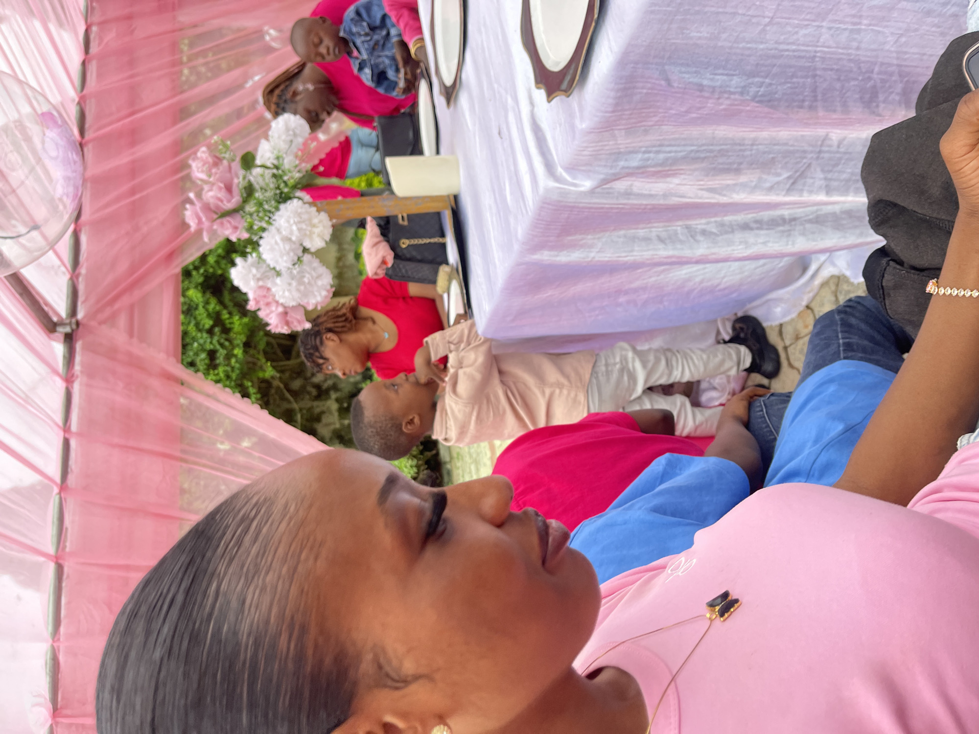 A gathering is taking place outdoors in Gura Top, Nigeria, under a pink draped canopy. In the foreground, a woman wearing a pink t-shirt and a butterfly pendant necklace is partially visible, her head turned to the side and looking down. To her right, a child in a light-colored shirt and white pants stands near a table. Several other people, including women and children, are seated or standing around the table, which is covered with a white and pink tablecloth and set with plates. A bouquet of pink and white flowers is placed on the table. The environment appears to be a garden or outdoor seating area with greenery visible in the background.