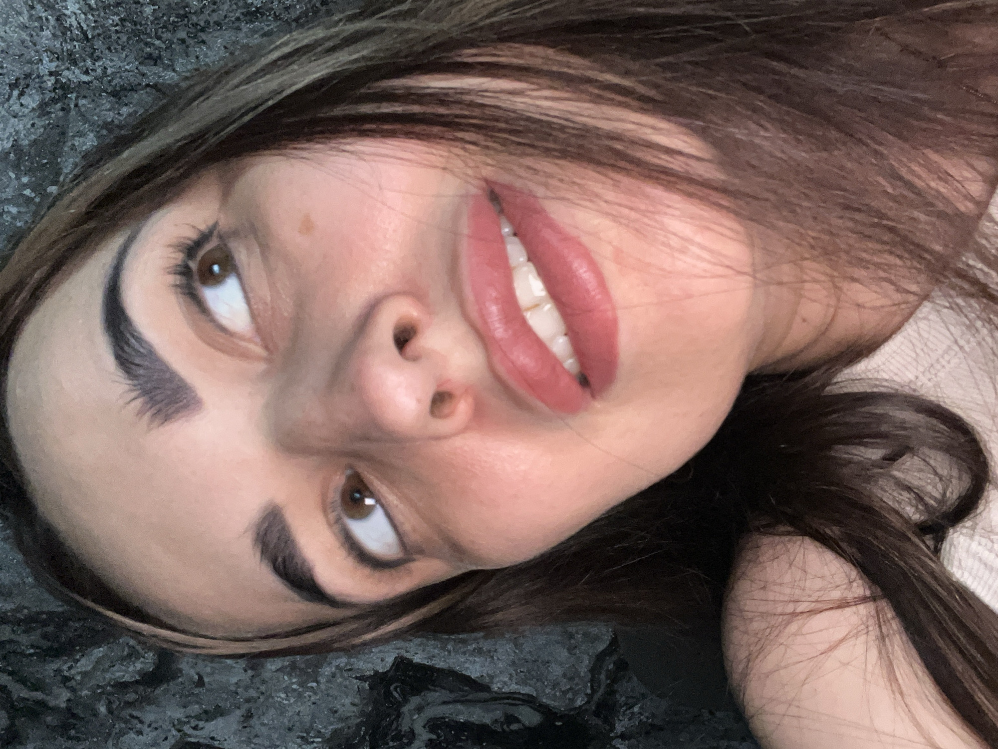 A close-up portrait shows a young woman captured from a slightly elevated and rotated perspective. Her head and upper torso are visible, with long, dark brown hair framing her face. She has prominent dark eyebrows and brown eyes that are looking upwards and slightly to her left. A small nose ring is present on her left nostril. Her lips, colored with a bright pink-red lipstick, are slightly parted, revealing her white teeth.
The woman appears to be lying down on a dark, mottled background that resembles stone or a textured wall, with shades of grey and black. A light-colored, possibly knitted or ribbed, garment can be seen on her right side (viewer's right). The lighting is soft and even, highlighting her features. Her expression is neutral, perhaps contemplative or subtly hinting at a smile. No text is visible in the image.