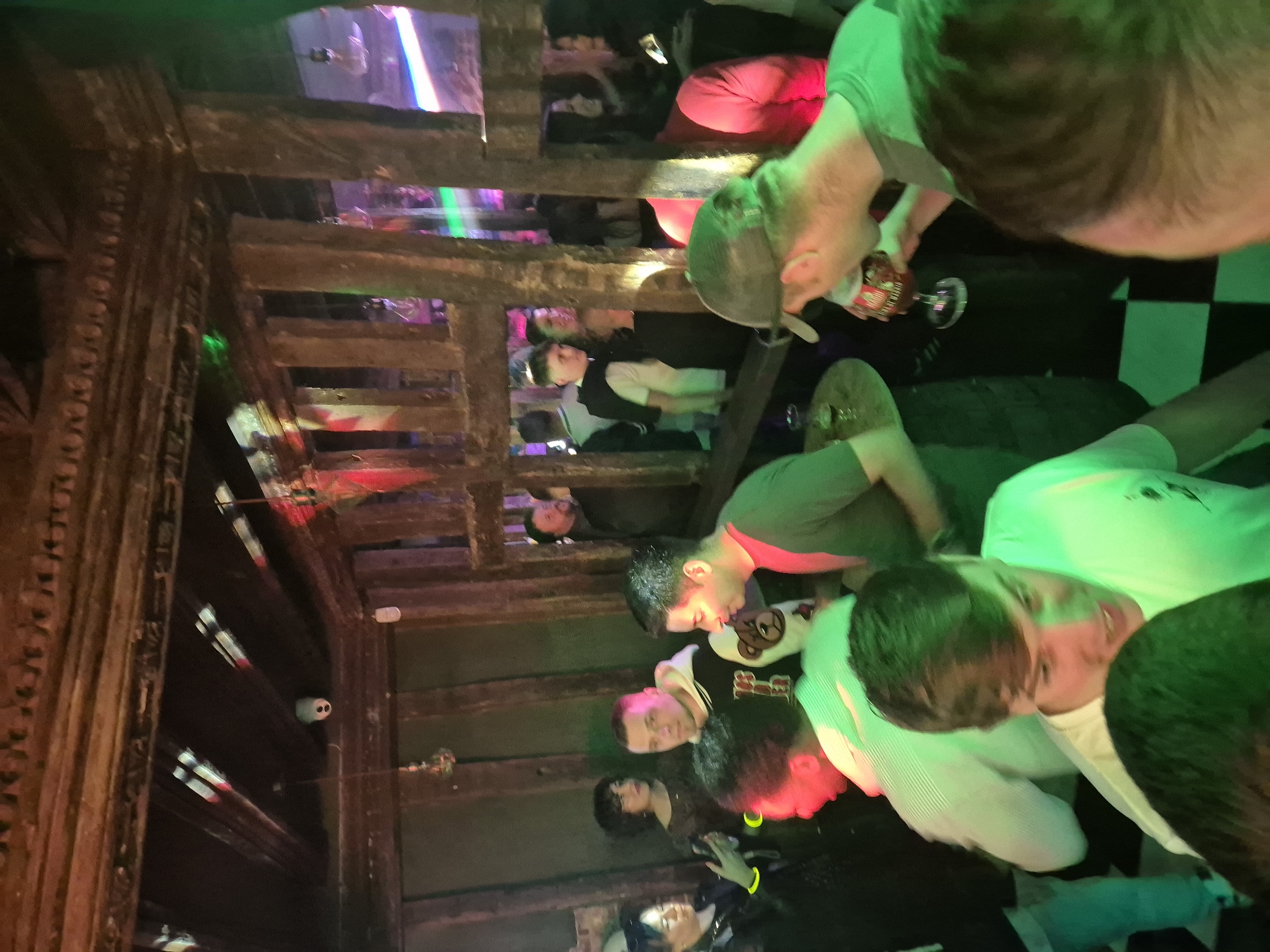 The image captures a crowded indoor space, possibly a multi-level bar or club, located in West Suffolk, United Kingdom. The camera angle is unusual, seemingly looking upward through a wooden latticework structure, possibly a stairway or decorative element. Multiple people are visible on different levels. Some are standing, others are seated. Individuals are dressed in a variety of casual attire. The environment appears dimly lit with colorful lighting effects in shades of red, green, and purple. Some people are holding drinks. There is limited visible interaction between people. A checkered floor pattern is visible in a small area.