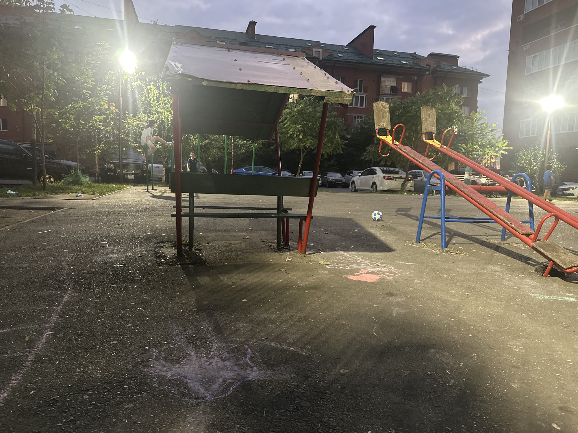 An outdoor children's playground, likely located in a residential area of Atabe, Georgia, is depicted. The scene is illuminated by artificial light sources, suggesting it is either dusk or night. Dominating the foreground and midground are playground equipment, including a seesaw with red metal framing and wooden seats, and a covered structure with a pitched roof supported by red metal poles and green framing. Chalk drawings are visible on the asphalt ground, with some appear to be hopscotch grids.
Several vehicles are parked in the background, including a white sedan and darker-colored cars. Residential buildings, characterized by brick facades and multi-story designs, are visible behind the playground. Trees with green foliage are interspersed throughout the scene.
Two individuals are present. One is a male child, appearing to be in his mid-to-late teens, actively engaged in exercising on a horizontal bar set up near the covered structure. He is wearing a white t-shirt and shorts. Another individual, a male child, stands near the covered structure observing. A soccer ball is positioned on the ground between the seesaw and the covered structure, partially in the shadow cast by the latter. The sky above is overcast with clouds.