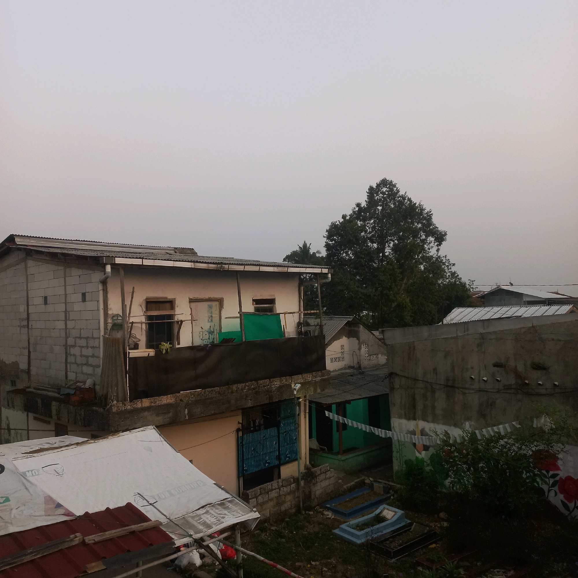 The image depicts an overcast sky over a residential area in the city of Bogor, Indonesia. In the foreground, a white tarp covers a slanted roof supported by wooden beams, with a red corrugated metal roof visible beneath it. To the left, a building constructed with cinder blocks is partially visible, with a corrugated metal roof.
The middle ground features a building with a white facade, a corrugated metal roof, and several windows. A dark tarp is draped over a section of this building. Next to it, a smaller building with a green facade and blue metal gates is visible. A wall with a mural depicting flowers separates this area from a section with several graves marked with blue surrounds.
In the background, a large tree with dense foliage dominates the right side of the frame. Beyond the trees and buildings, the sky is a uniform light gray, suggesting overcast conditions or perhaps early morning or late evening light. The overall scene conveys a sense of a densely populated urban environment.