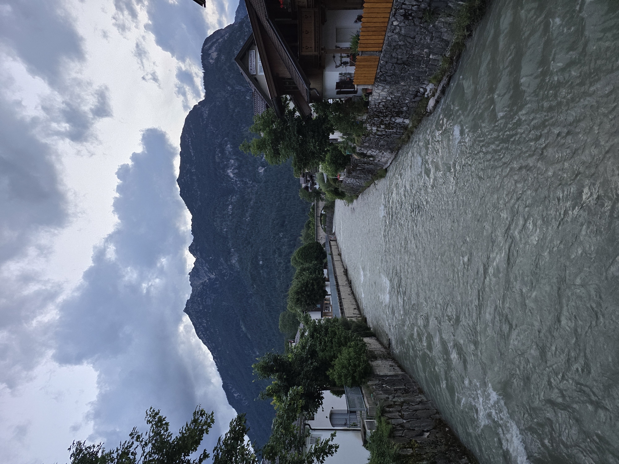The image depicts a wide, murky river flowing through a European town. The riverbanks are lined with stone walls and interspersed with trees and buildings. On the right bank, a multi-story building with dark roofing and wooden balconies is visible. A bright orange wooden fence is also present. On the left bank, a white building with a grey door and shutters is seen, with trees and greenery surrounding it. In the background, a lush green mountain range dominates the skyline under a cloudy sky. A stone bridge can be seen in the distance, spanning the river. The scene suggests a tranquil, possibly evening setting in a mountainous town. Without further information, the specific location within Garmisch-Partenkirchen, Germany cannot be definitively identified.