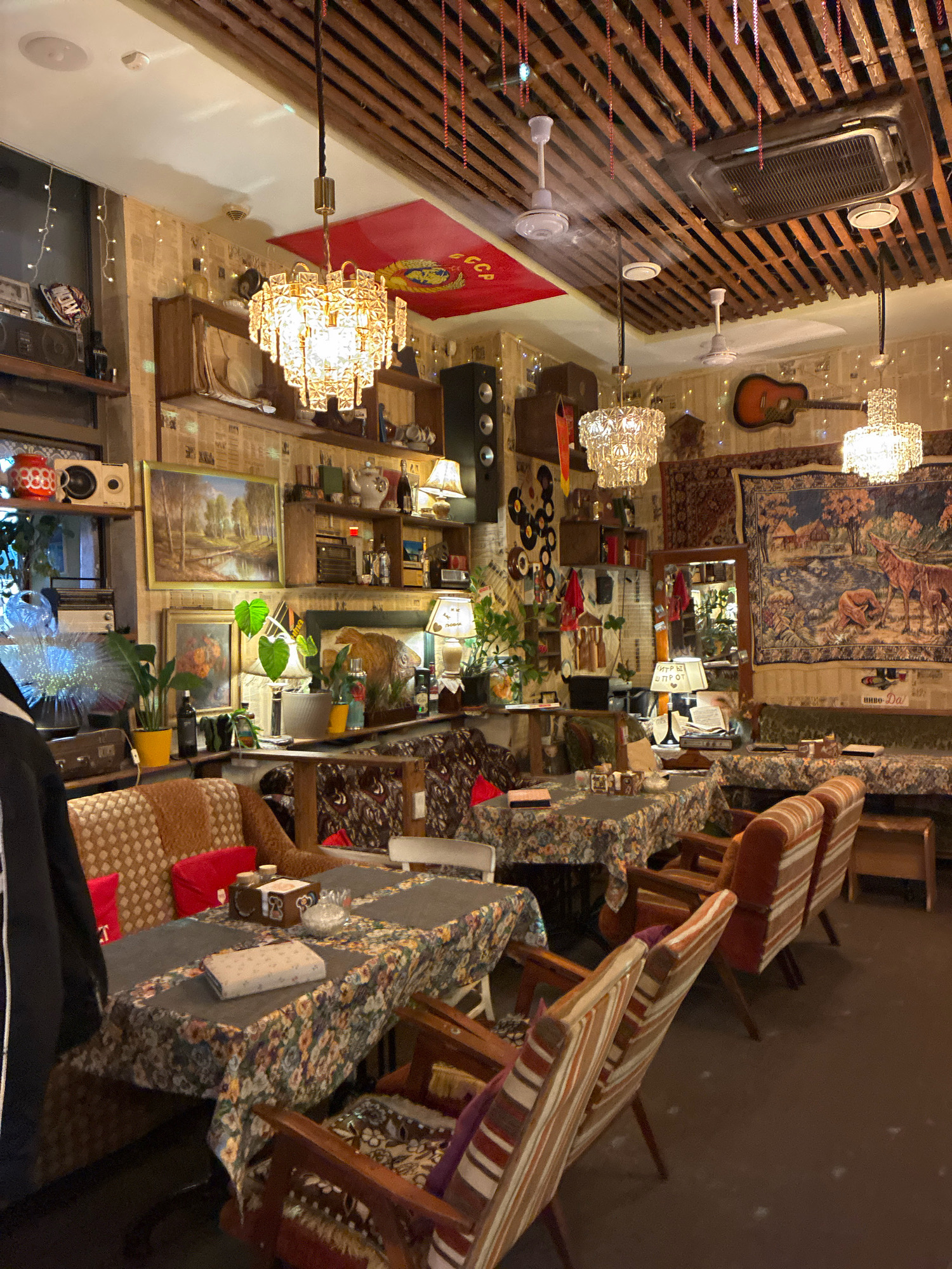 The image depicts the interior of a dimly lit establishment, likely a cafe or restaurant, characterized by its vintage and eclectic decor. Suspended from a slatted wooden ceiling, several elaborate crystal chandeliers illuminate the space, casting a warm glow. Two ceiling fans are visible, one on the left and another further back, along with an air conditioning unit. A red flag with a yellow emblem, possibly a Soviet-era symbol, hangs prominently on the back wall.
The walls are covered in what appears to be newspaper clippings, adding to the retro aesthetic. Shelves are filled with various objects, including framed pictures, vintage electronics like radios and record players, bottles, and decorative items. A guitar is mounted on the wall behind a tapestry depicting a forest scene with deer.
Several tables are set with floral-patterned tablecloths. One table in the foreground is set with a folded cloth, a wooden box containing condiments, and a small glass jar. Chairs surrounding the tables vary in style, with some featuring patterned upholstery and others being more simply wooden.
In the foreground, on the left, a dark-colored coat is draped over a chair. The overall atmosphere suggests a nostalgic and perhaps bohemian ambiance.
This establishment is identified as the "Stolichny" bar, located at 8 Ulitsa Ordzhonikidze in Sochi, Russia.